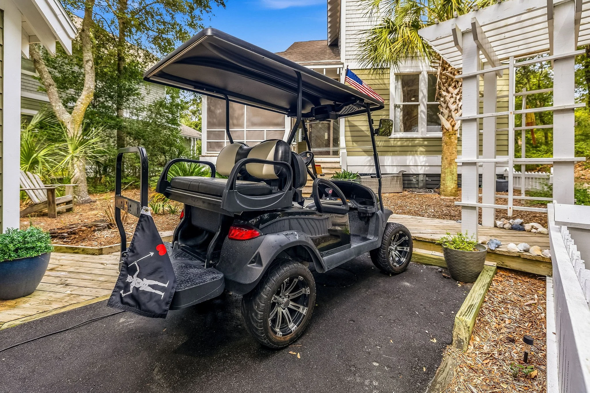 A black golf cart parked on a driveway with a golf club and bag in front of it, outside a house with beige and white exterior, surrounded by trees and plants.