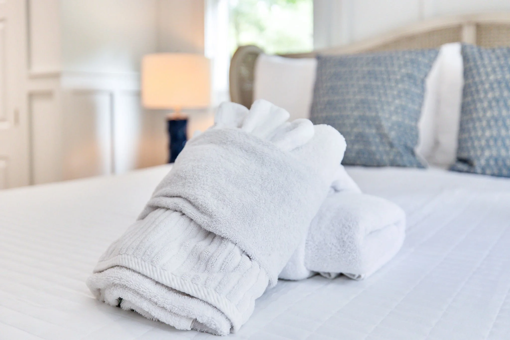 A neatly folded white towel on a bed with white sheets, with blue patterned pillowcases and a white pillow in a bright, softly lit bedroom.
