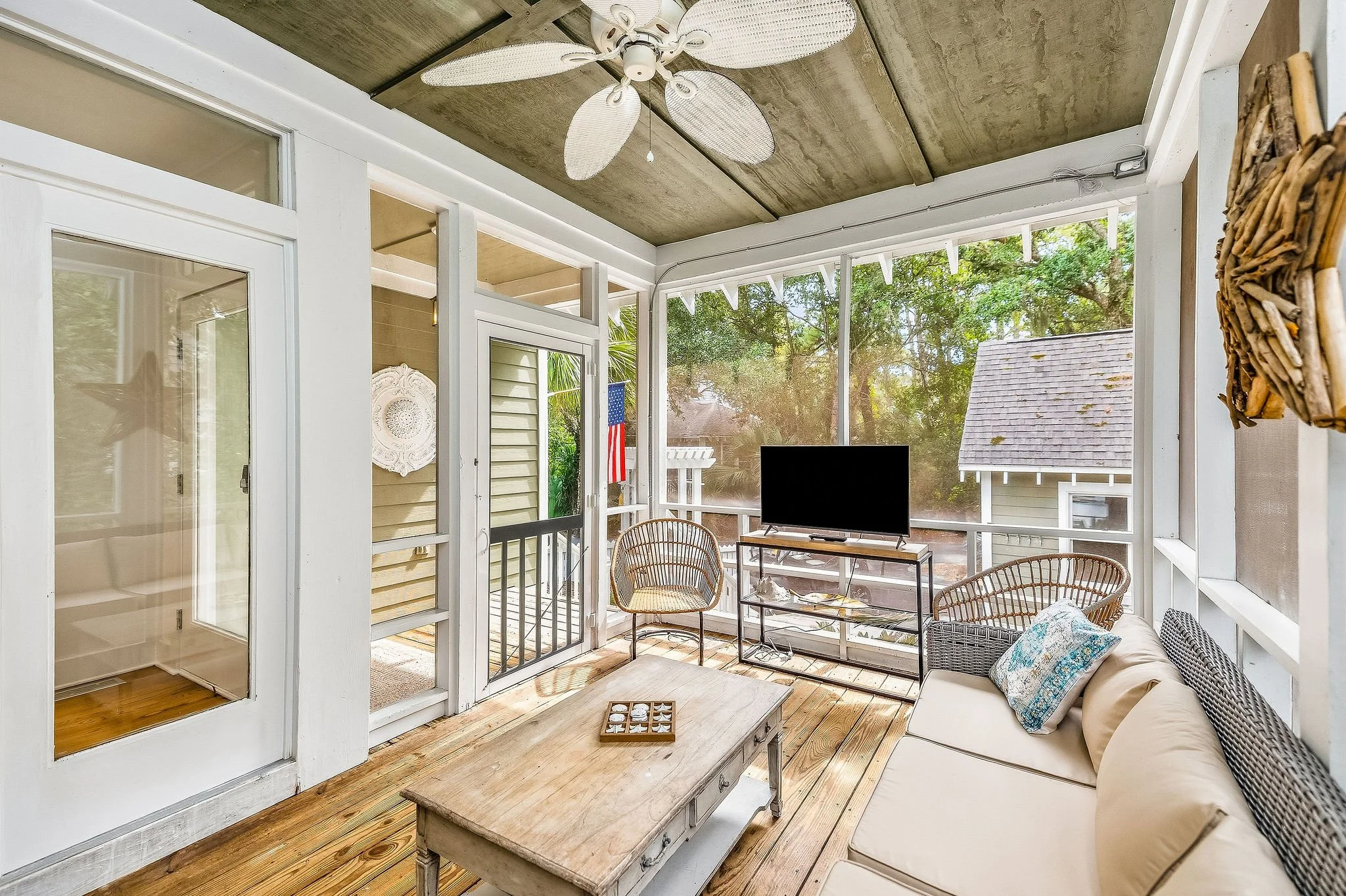 A sunroom with screened windows, outdoor furniture including wicker chairs and a couch, a television on a stand, a ceiling fan with palm leaf-shaped blades, and a wooden coffee table.