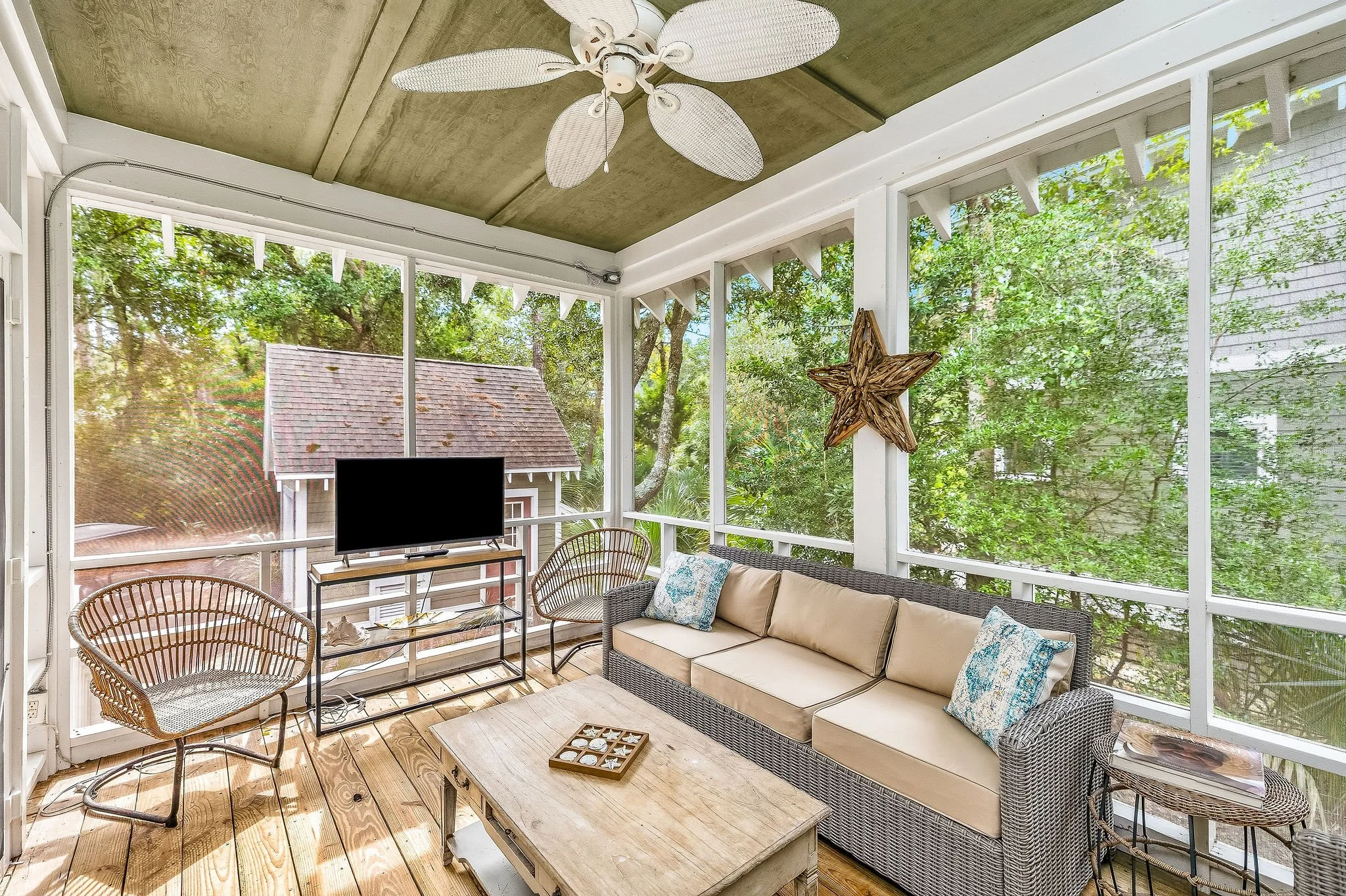 A screened-in porch with wicker chairs, a beige sofa with patterned pillows, a wooden coffee table, a television on a small stand, and trees visible through the screens.