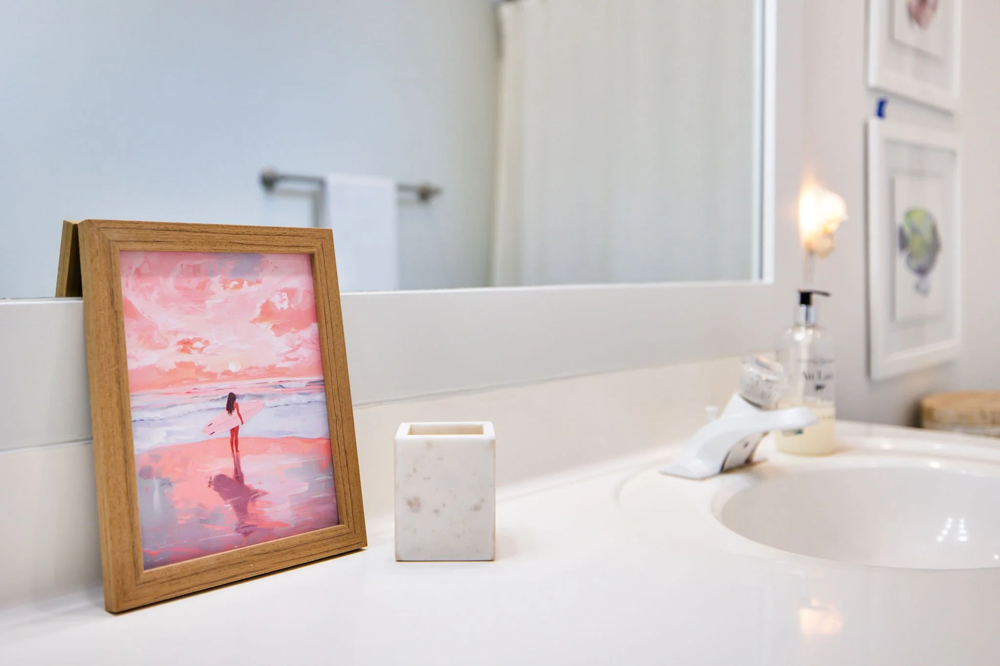 A bathroom countertop with a framed pink and orange watercolor painting of a person holding a pink umbrella, a marble container, and a soap dispenser. In the background, a mirror, a towel rack, and framed artwork are visible.