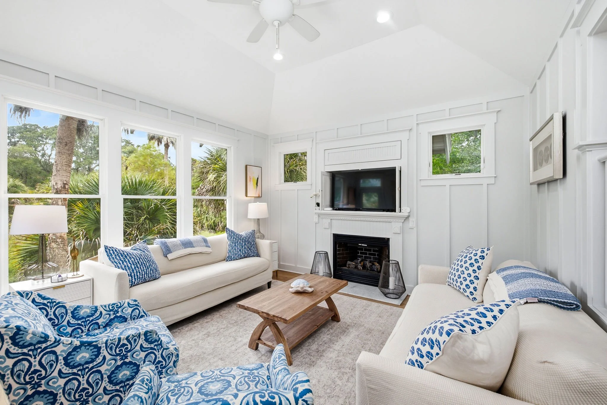 Living room with white walls, large windows, and a white ceiling fan. It features a fireplace, flat-screen TV, and furniture including white sofas with blue patterned pillows and a wooden coffee table. There's lush greenery visible outside the window