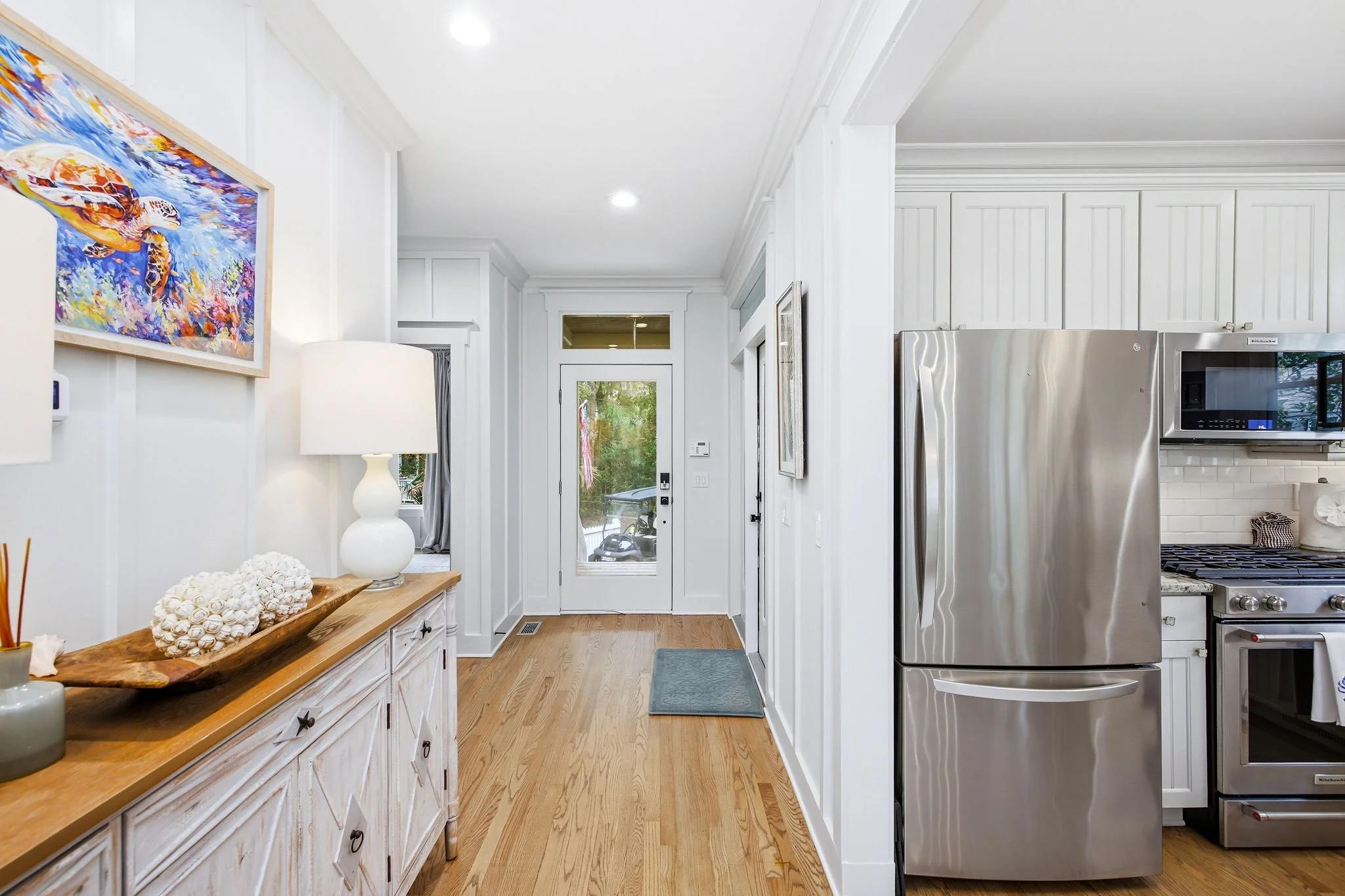 The image shows a bright, modern kitchen with white cabinetry, stainless steel appliances including a refrigerator and oven, a wooden console with decorative shells and a white lamp, hardwood floors, and a view of the outdoors through the door.