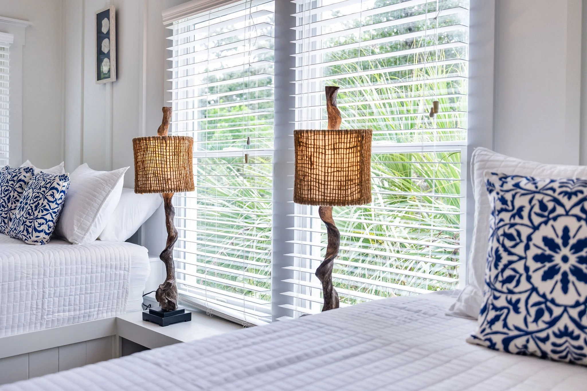 A bright bedroom with white walls and large windows with white blinds. There are two straw table lamps with curved wooden bases on a white side table. Part of a bed with white quilted bedding and blue and white patterned pillows is visible.