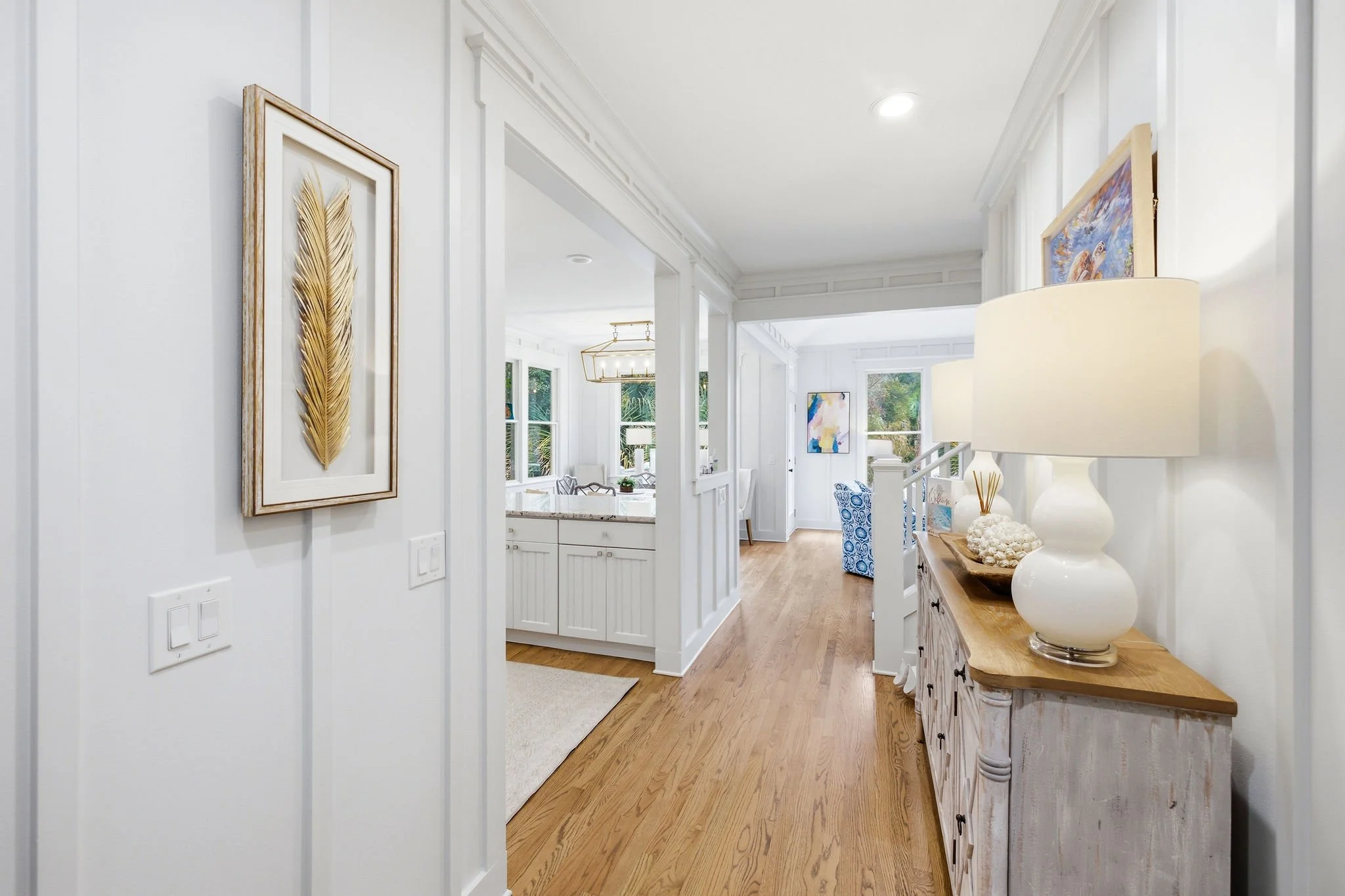 The image shows a bright, spacious interior hallway of a house with white walls and wooden floors. Artworks are mounted on the walls, including a framed piece of a golden feather on the left. To the right, a wooden console table holds a large white l