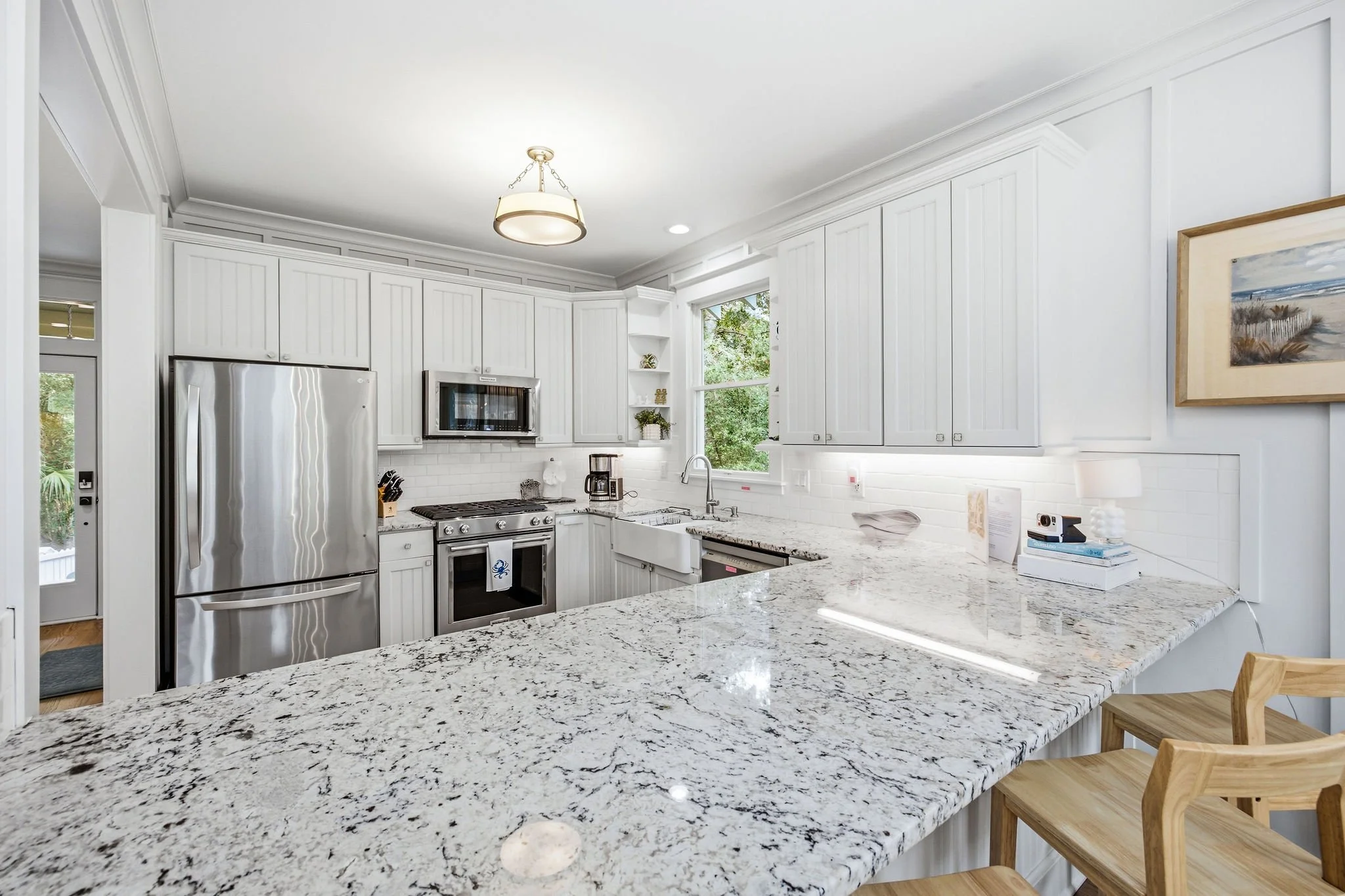 A modern white kitchen with granite countertops, stainless steel appliances, white cabinets, and a small table with chairs. There are windows letting in natural light and a painting on the wall.