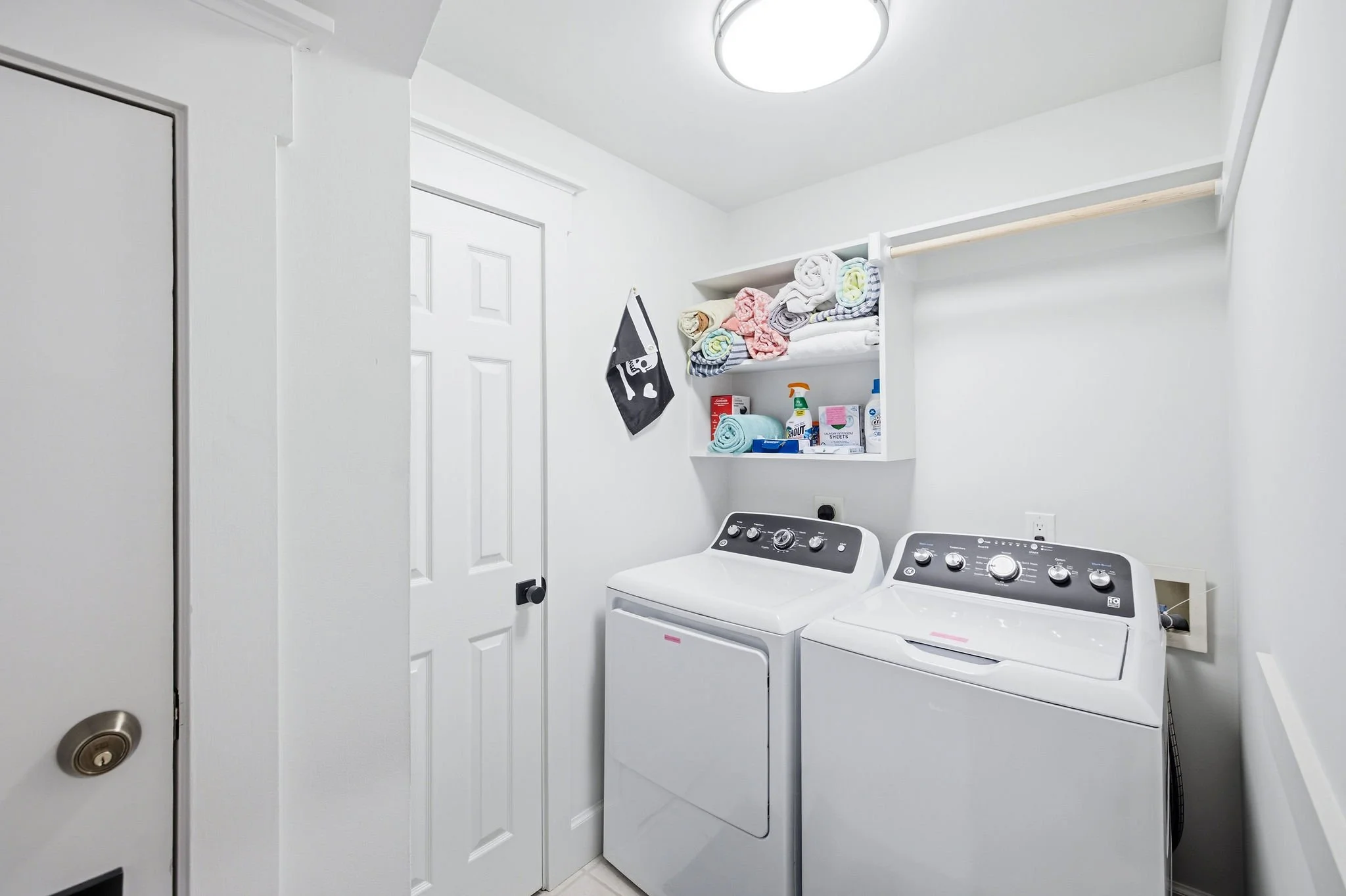 A laundry room with white walls, a white ceiling, and a white door. Two white washing machines are side by side under a shelf that holds folded towels and laundry supplies. An overhead light fixture illuminates the room.