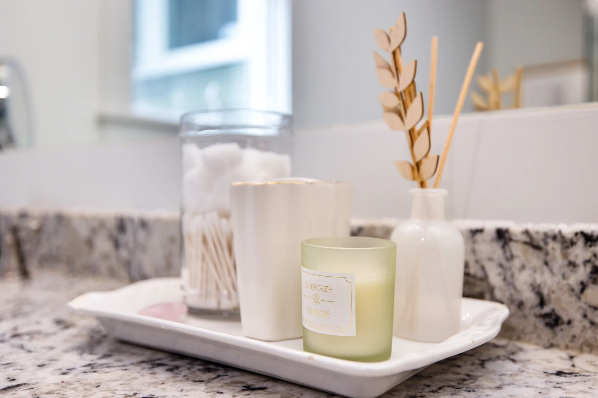 A white tray holding a green candle labeled 'Energize,' a white candle, a glass container with cotton balls, and a container with wooden sticks with decorative leaves.