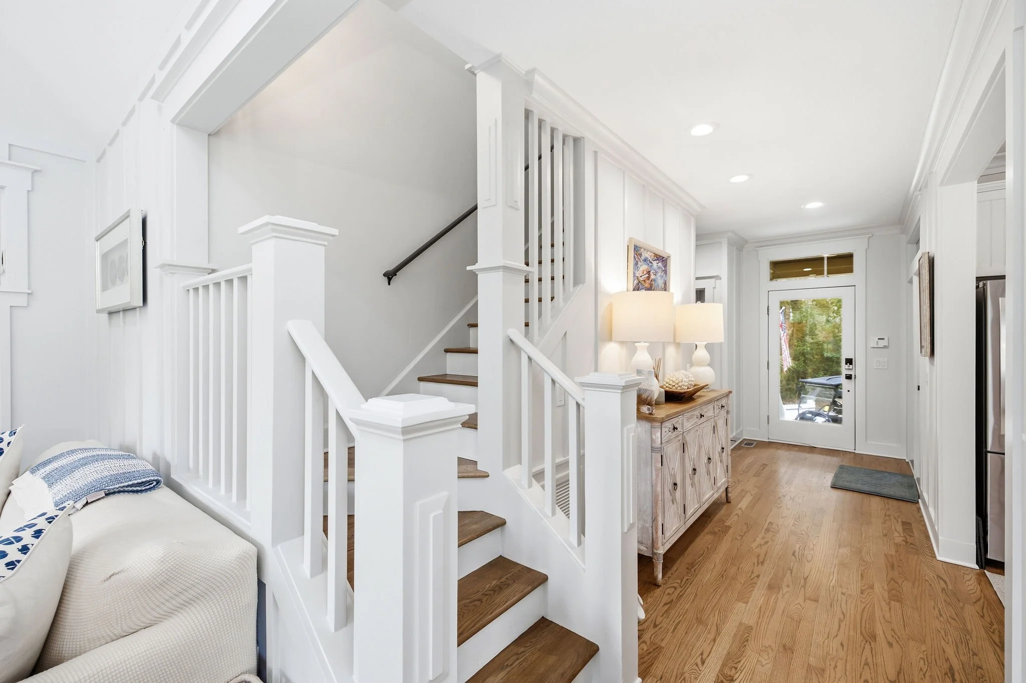 Interior view of a modern home entryway with white walls, wooden flooring, a staircase with white railings and wooden steps, and a wooden console table with decorative lamps and artwork, near a glass door.