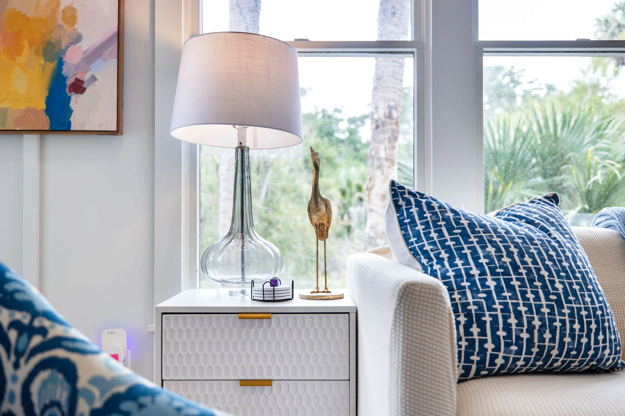 A bright living room with large windows showing trees outside. There is a white side table with a glass lamp, a small figurine, and a striped box. A beige armchair and a sofa with blue and white patterned pillows are partially visible. A colorful abs