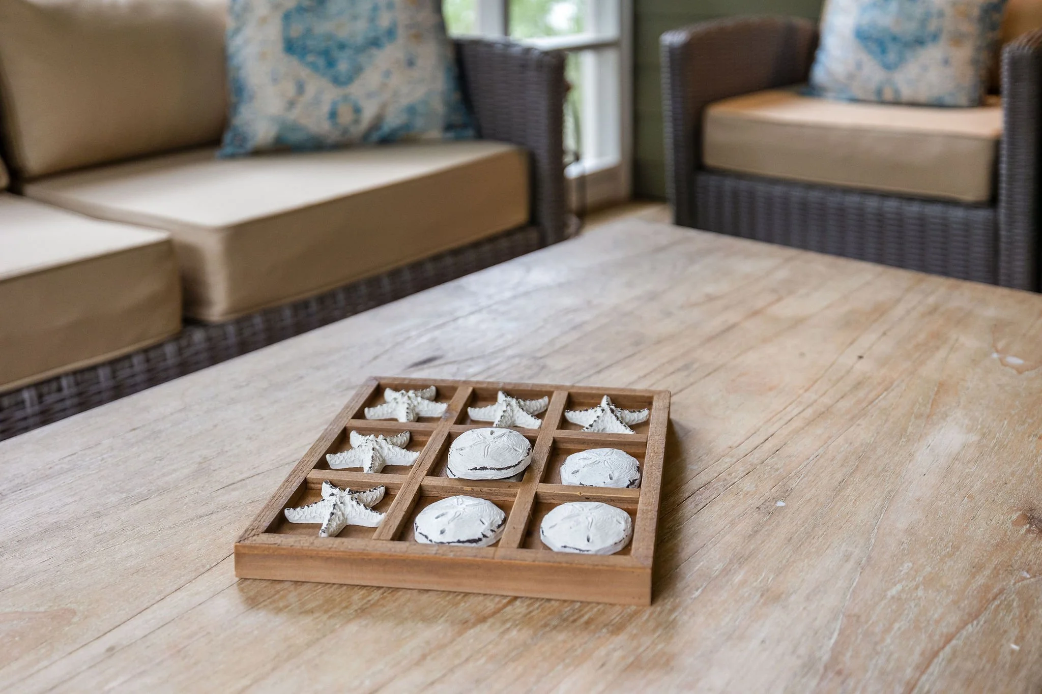 A wooden tray with several white seashell-shaped cookies and round cookies on a wooden table in a cozy room with wicker and upholstered furniture, and a window in the background.