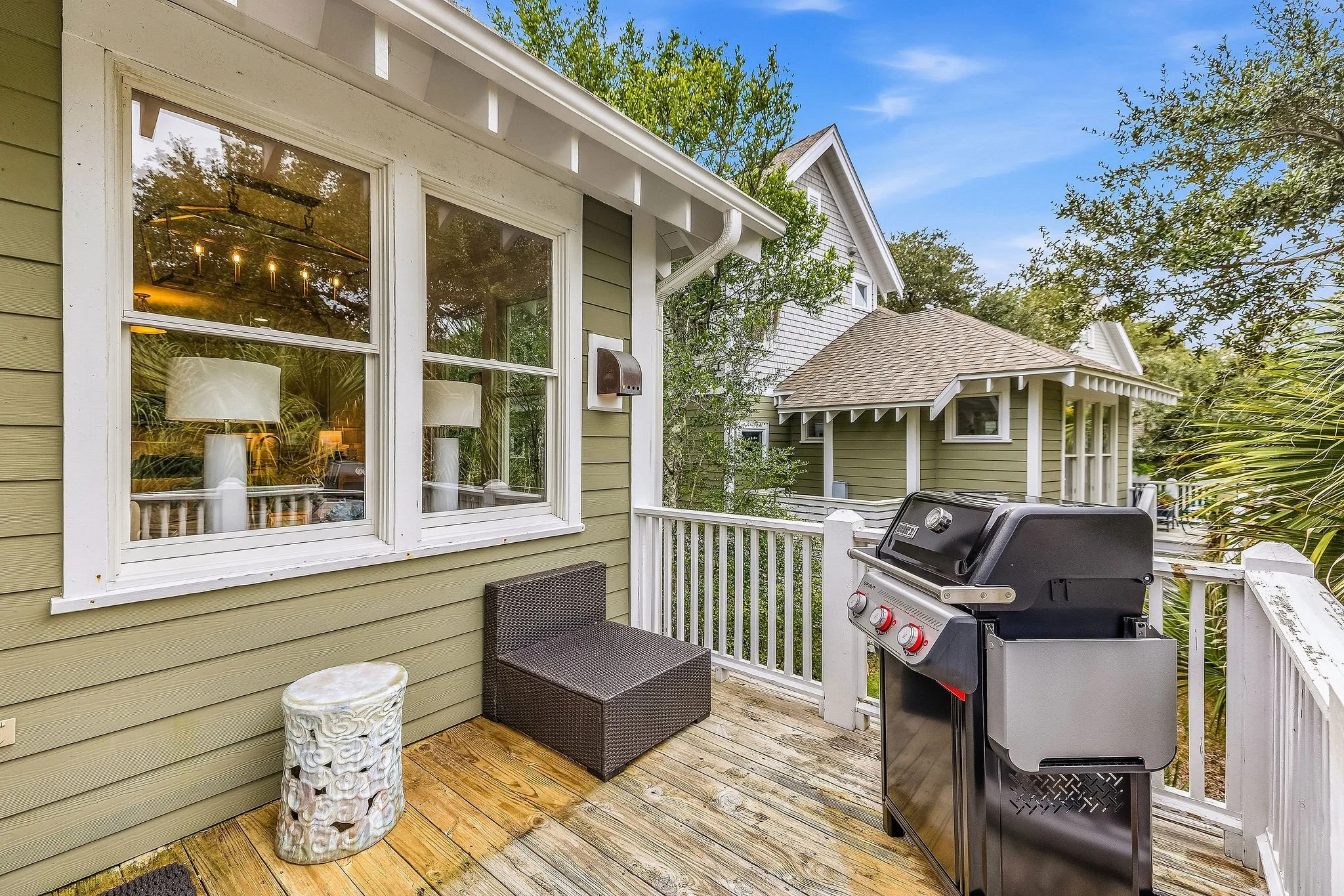 A backyard patio with a green house, a TV, a wicker chair, a decorative white side table, and a barbecue grill on a wooden deck, surrounded by trees and neighboring houses.