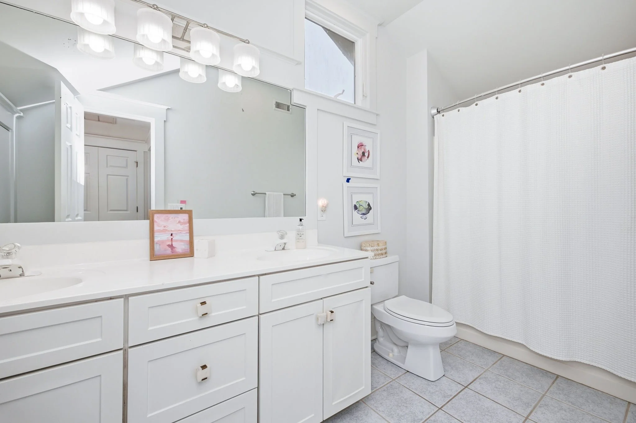 A modern white bathroom with a large mirror above a white vanity, featuring framed artwork of fish and flamingos on the wall, a white toilet, and a white shower curtain. The room has a high ceiling with a set of white lamp fixtures and a skylight.