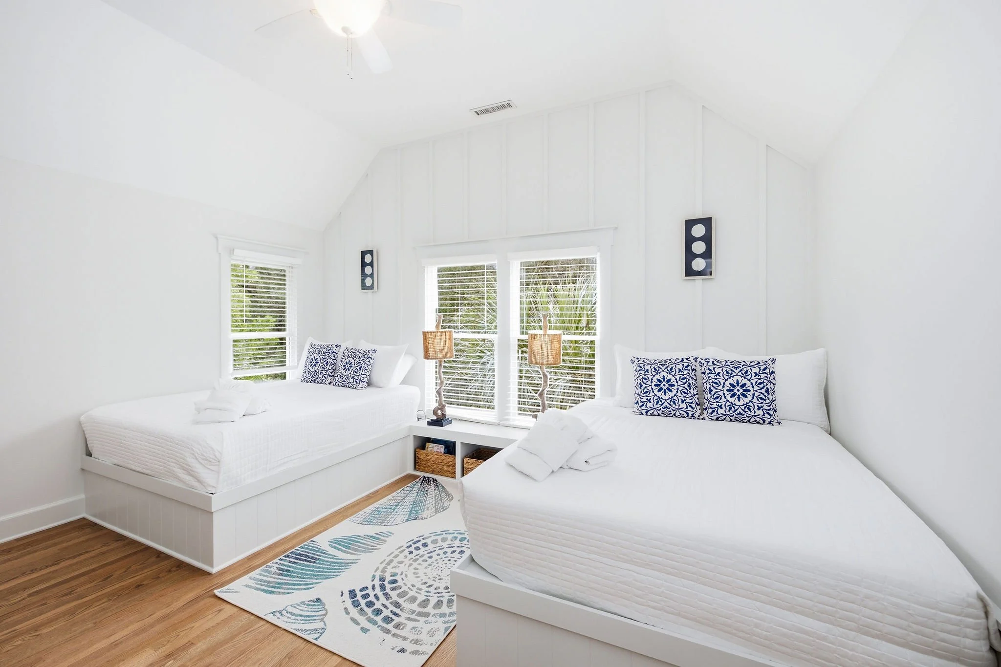 A bright white bedroom with two beds with white linens and blue patterned pillows, a window with white blinds, and wicker lamps on nightstands, with natural light coming in.