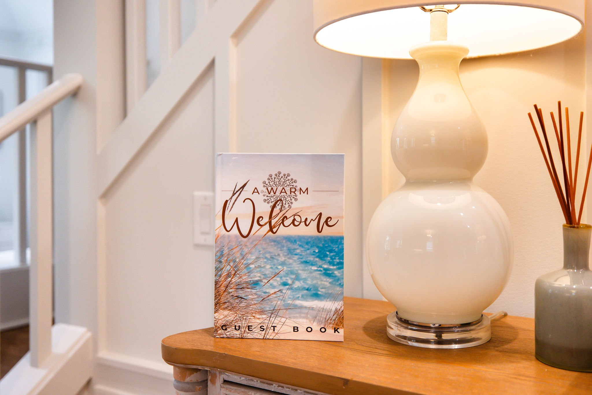 A bedside table with a large white ceramic vase lamp, a guest book with a beach scene cover, and a reed diffuser, inside a well-lit room.