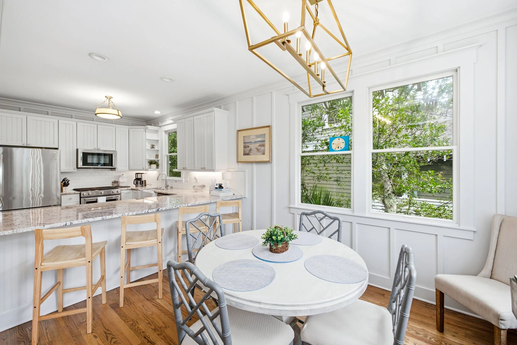 Bright kitchen and dining area with large windows, white cabinets, stainless steel appliances, and a round dining table with gray chairs and a small potted plant.