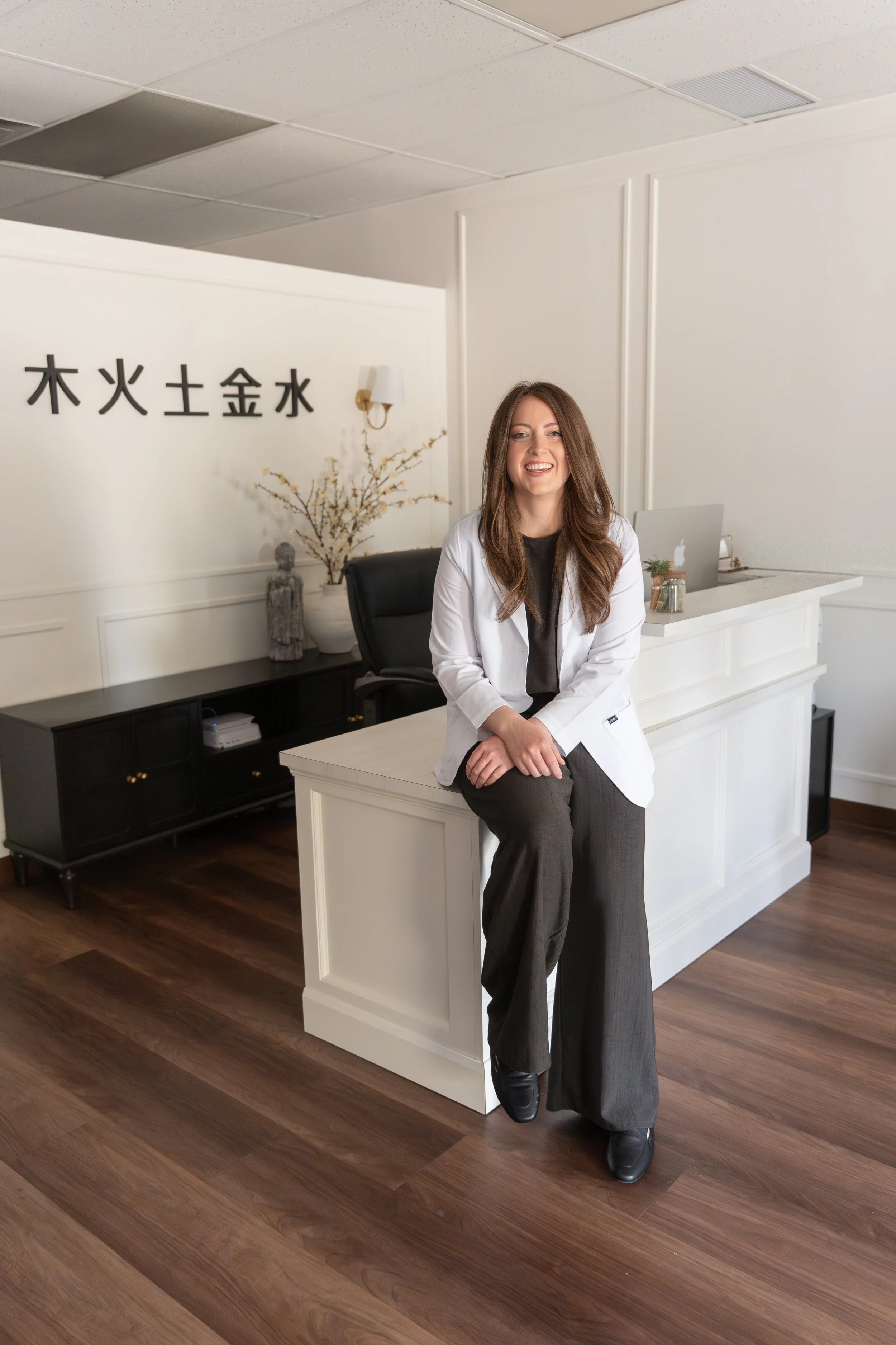 A female traditional chinese medicine practitioner sitting on a white reception desk in a modern office with white walls, wooden flooring, and decorative plants, smiling at the camera in Lacombe Alberta