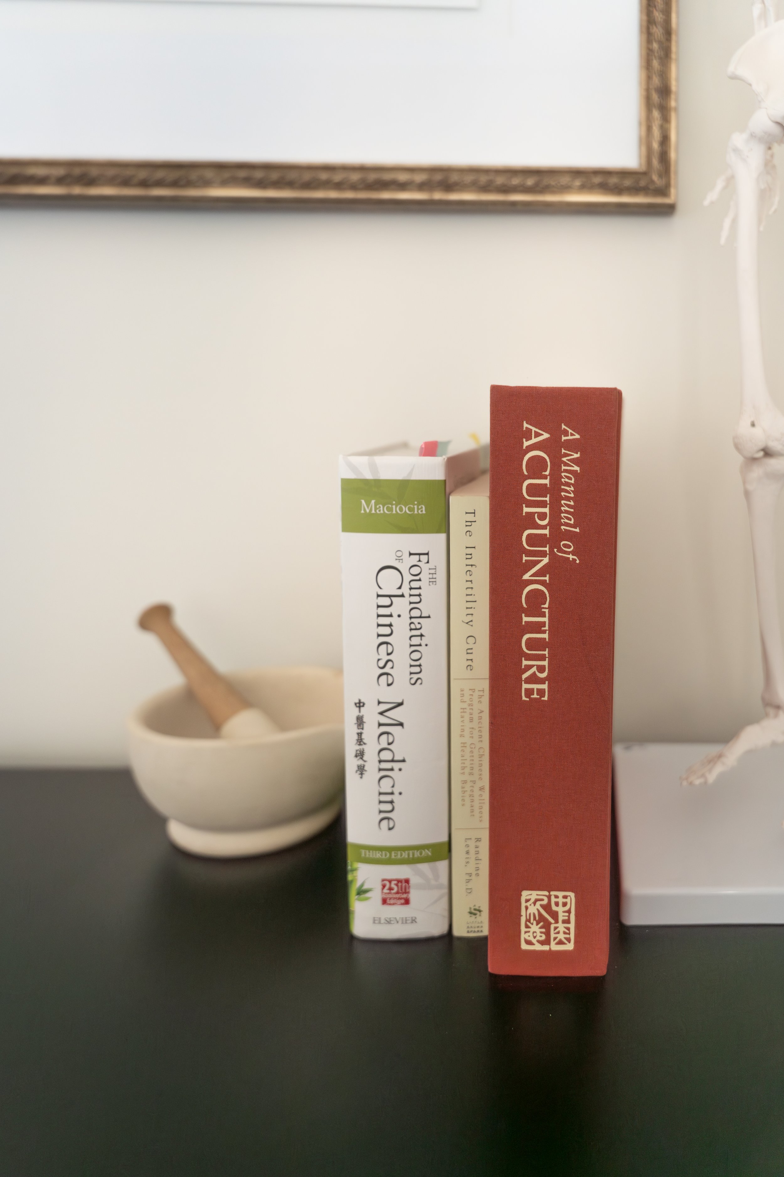 Three books standing upright on a black surface, with a small white bowl with a pestle behind them. The books are titled 'Maciocia', 'The Foundations of Chinese Medicine', and 'A Mayummu of Acupuncture'. A framed mirror is hanging on the wall above, and part of a skeletal model is visible on the right.