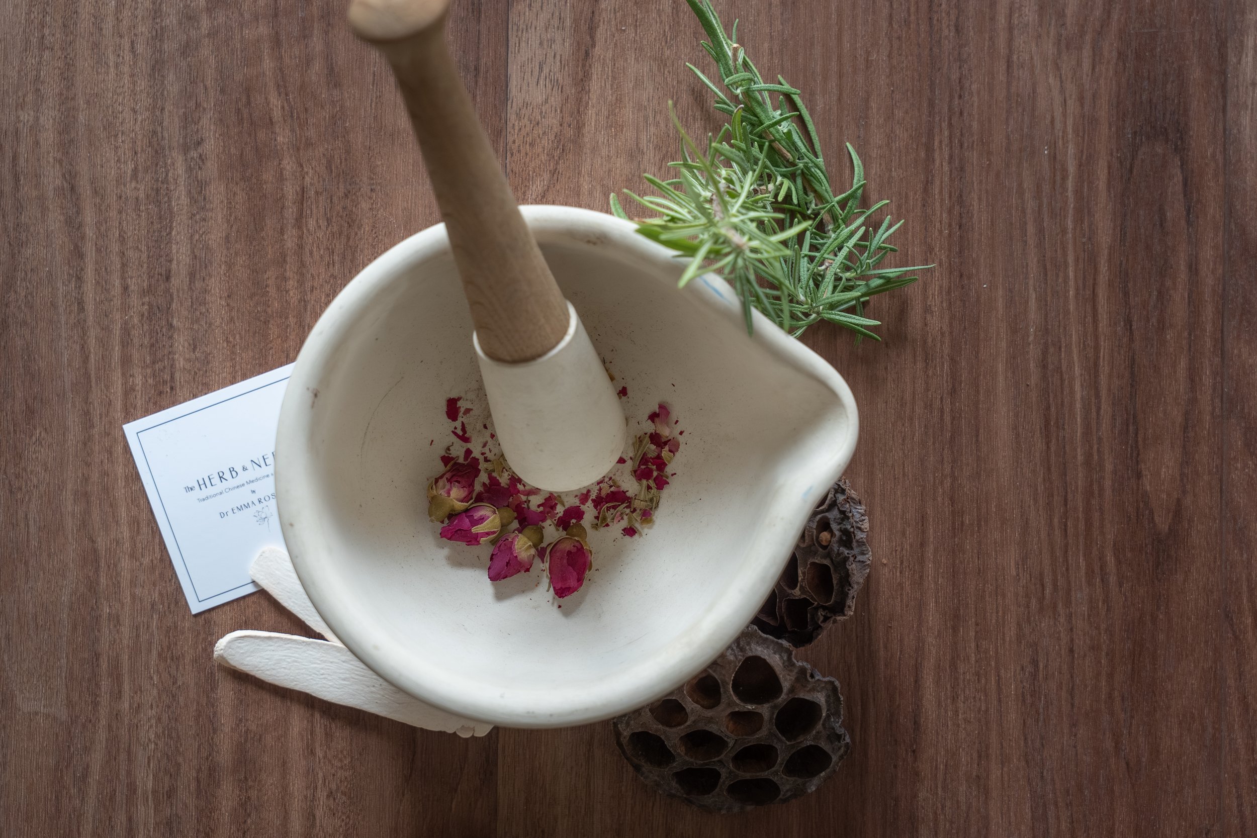 A ceramic mortar and pestle with dried rose petals inside, placed on a wooden surface next to a green herb plant, a piece of wood, and an informational card for Traditional Chinese Medicine clinic in Lacombe Alberta