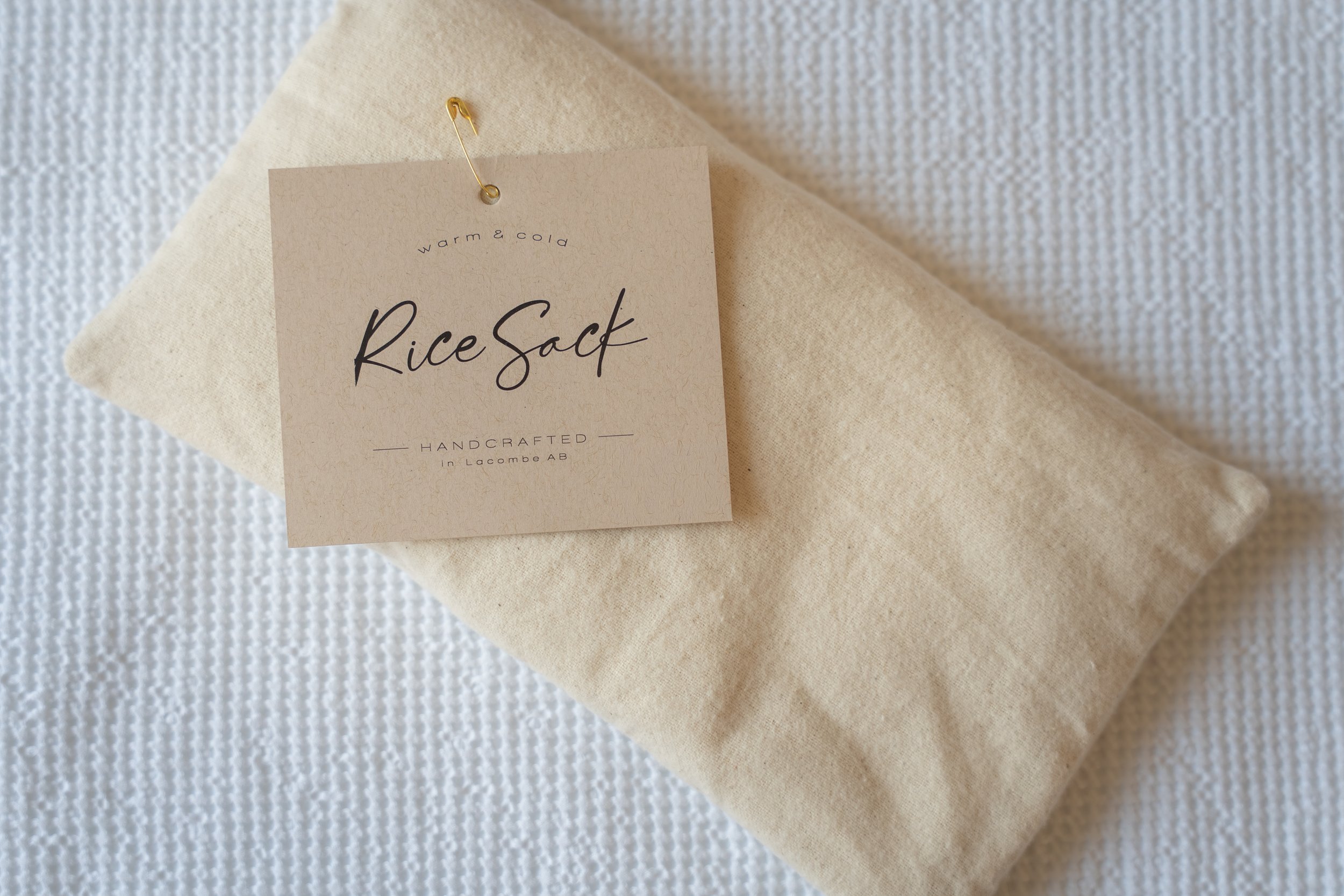 A beige fabric rice sack with a paper label attached by a gold pin, labeled 'Rice Sack' with the words 'warm & cold' and 'handcrafted in Lacombe AB', resting on a white textured surface.