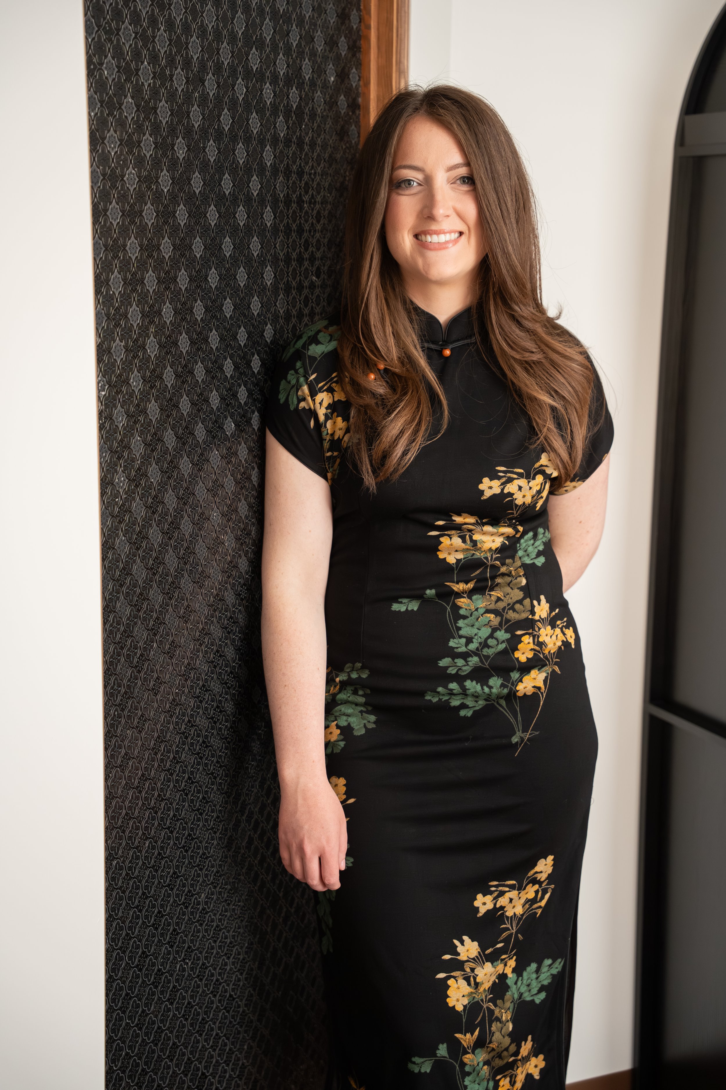 A female traditional chinese medicine practitioner with long brown hair smiling, wearing a black dress with yellow and green floral pattern, standing in an indoor setting near a decorative black wall and a doorway.