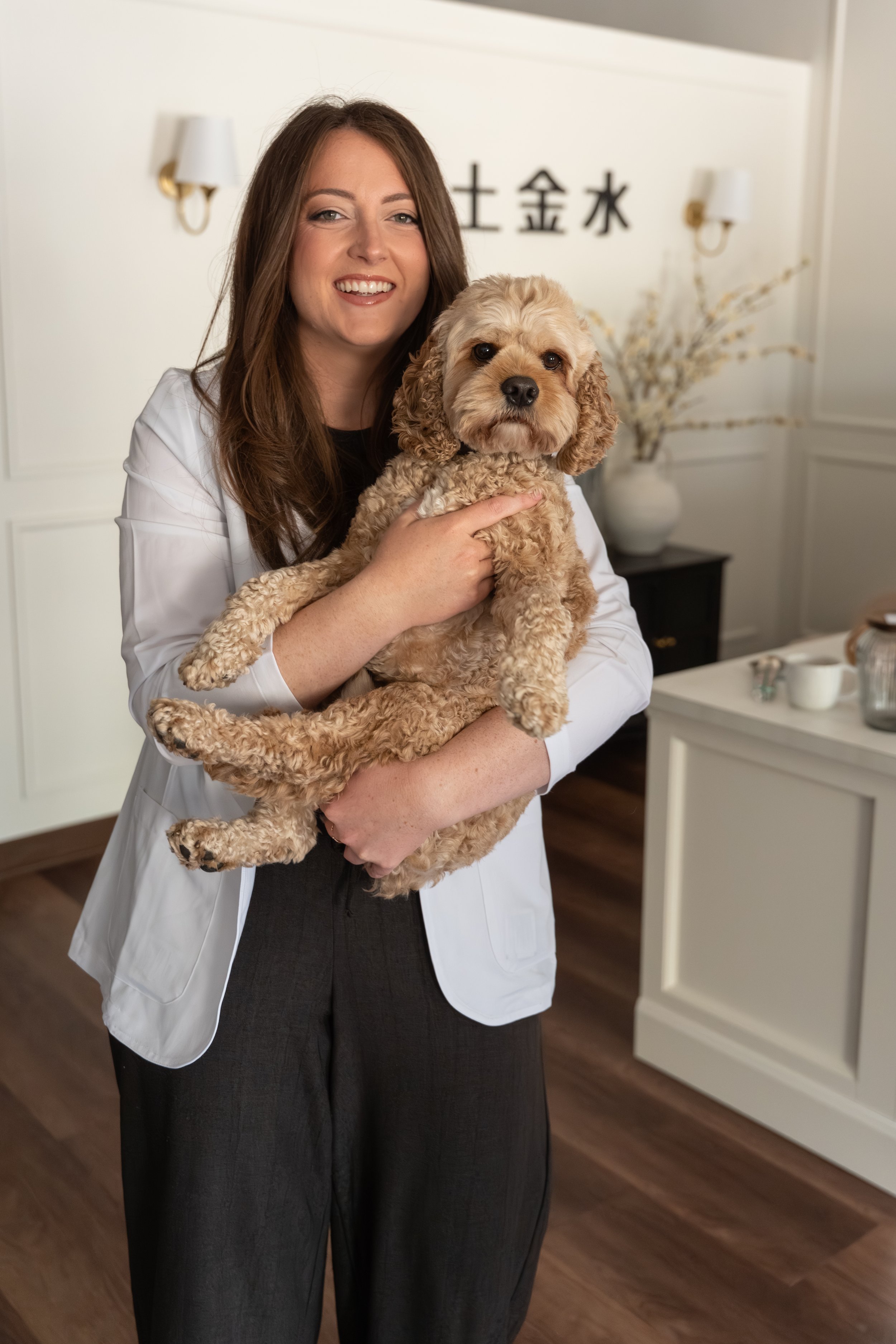 A female traditional chinese medicine practitioner with long brown hair holding a light tan curly-haired dog in an indoor room with white walls and dark wooden floors. There are two wall-mounted lamps and Japanese characters on the wall behind her.