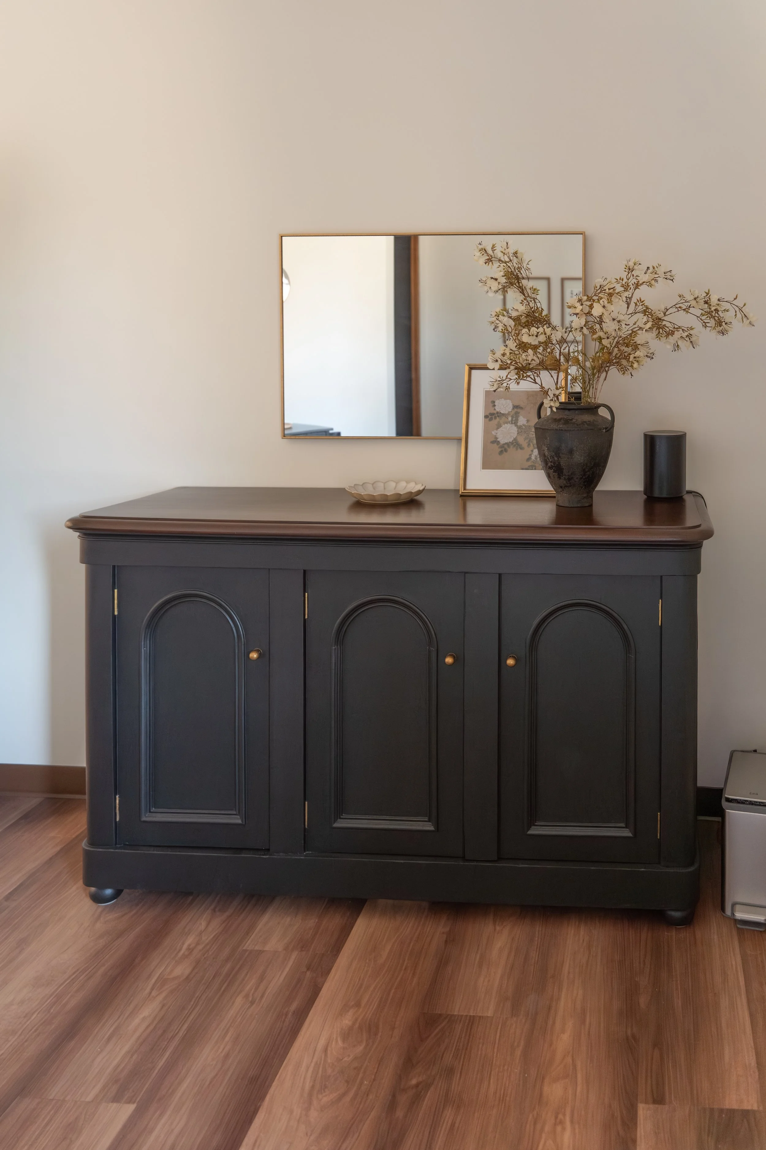 A dark blue wooden sideboard with gold accents and rounded panel doors, standing on a hardwood floor. On top of the sideboard are a framed floral artwork, a gray ceramic vase with dried beige flower branches, a small ceramic bowl, and a black electronic device. A mirror is mounted on the wall above the sideboard, reflecting parts of the room.