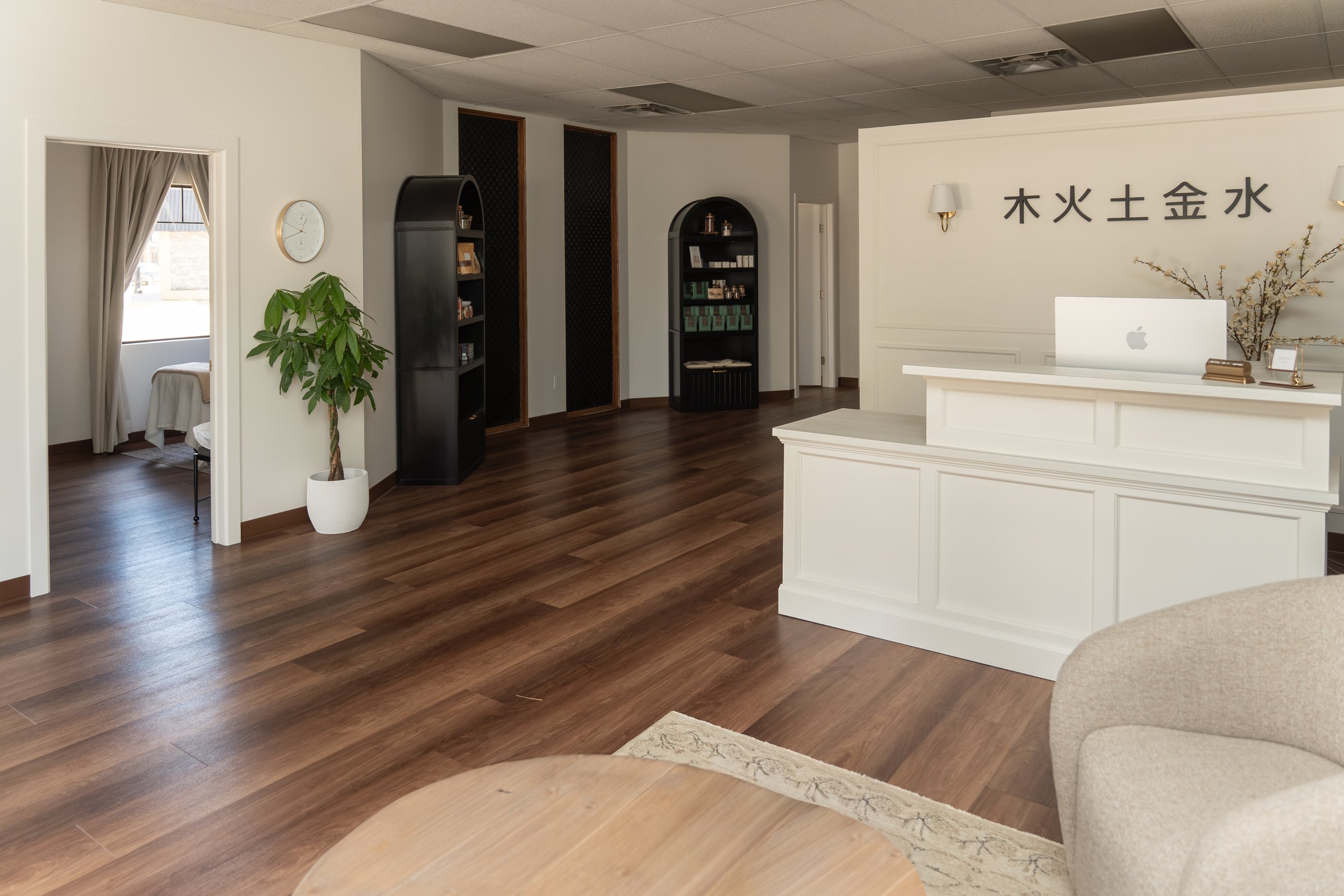 A modern reception area for Traditional Chinese Medicine clinic with a white desk, Apple computer, decorative plants, bookshelves, a wall clock, and Asian characters on the wall.