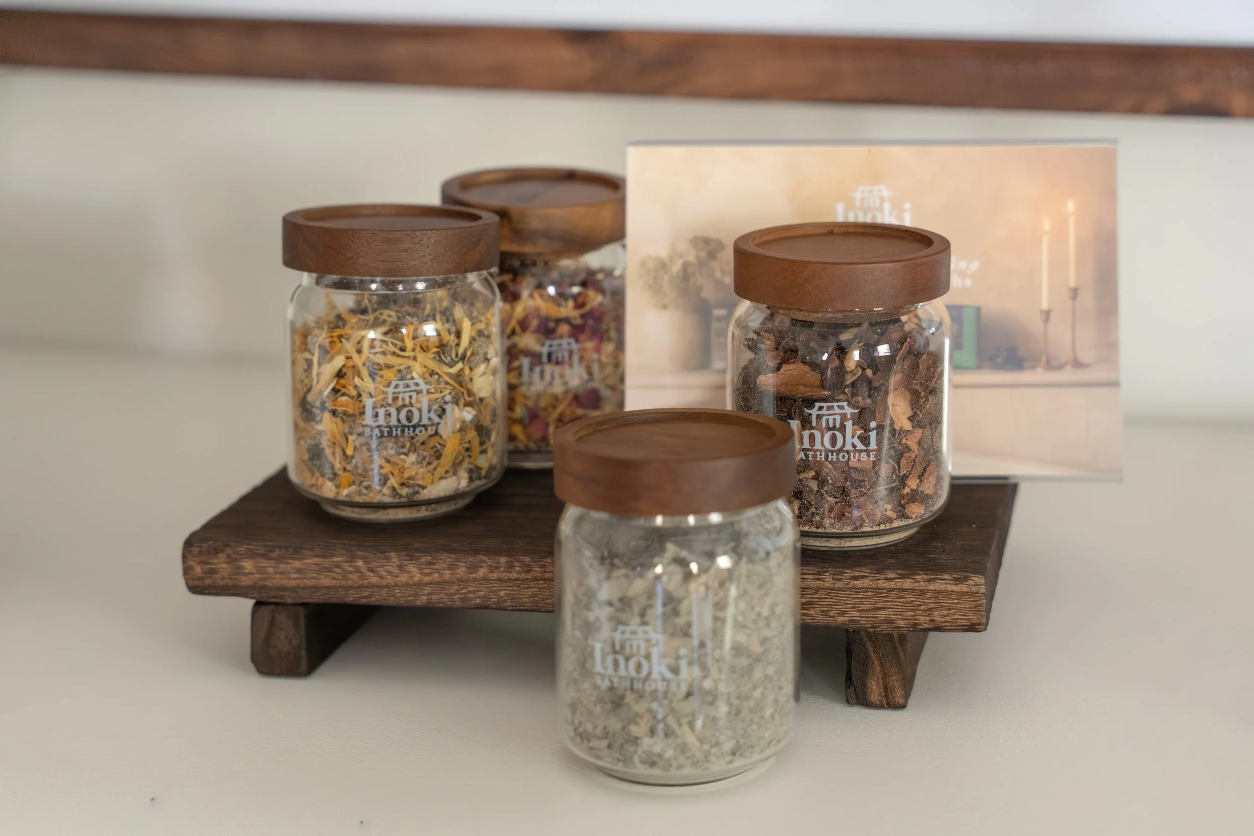 Four glass jars with wooden lids containing dried herbs, flowers, and spices are displayed on a wooden shelf with a white background in Lacombe Alberta.