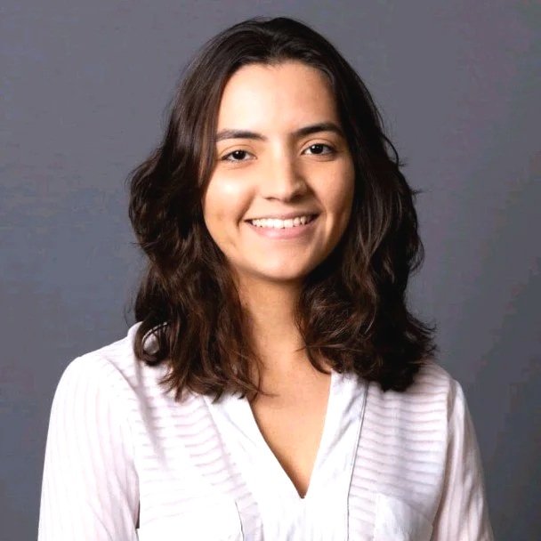 Retrato de uma mulher jovem sorrindo, cabelo castanho e vestido branco, contra fundo cinza.
