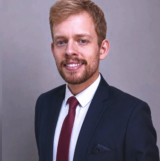 Homem sorrindo com cabelo castanho claro, barba e traje formal, usando blazer escuro, camisa branca e gravata vinho, em fundo cinza.