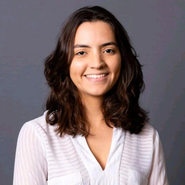 Retrato de uma jovem mulher com cabelo castanho escuro, pele clara e sorrindo, usando blusa branca com detalhes em relevo, contra fundo cinza.