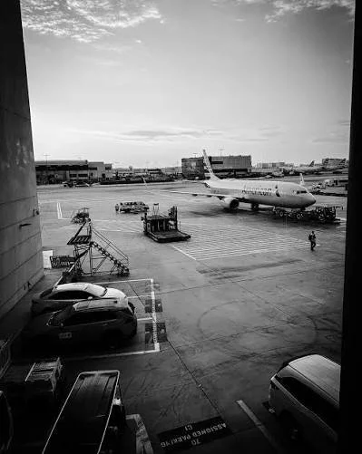 Trem de pouso de avião no aeroporto, estacionamento com carros, escada e pessoas ao ar livre, céu com algumas nuvens.