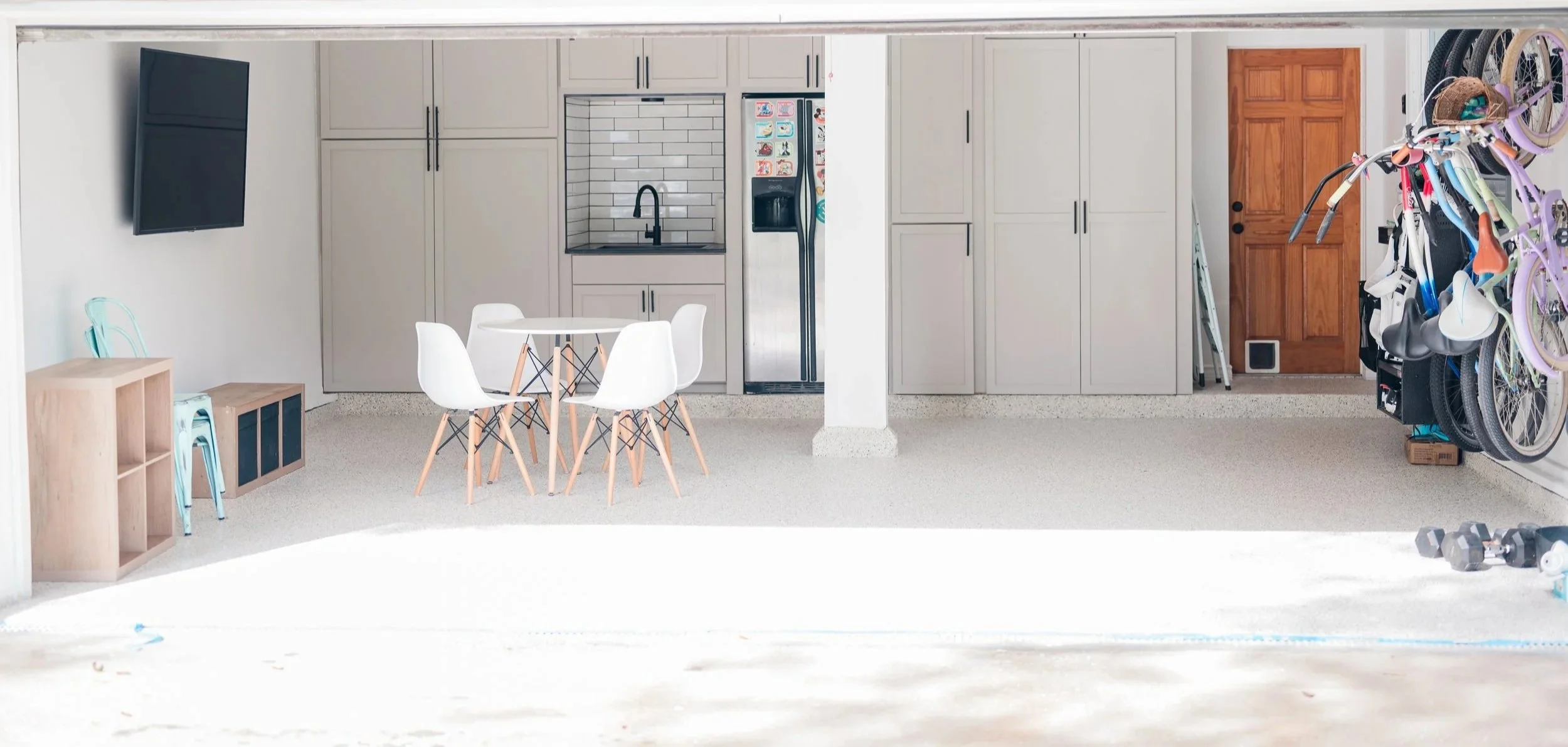 An indoor space that appears to be a garage or multipurpose room, with a kitchen area including cabinets, a dining table with four white chairs, and a wall-mounted television. On the right side, there are multiple bicycles, some with training wheels, and a pair of dumbbells on the floor.
