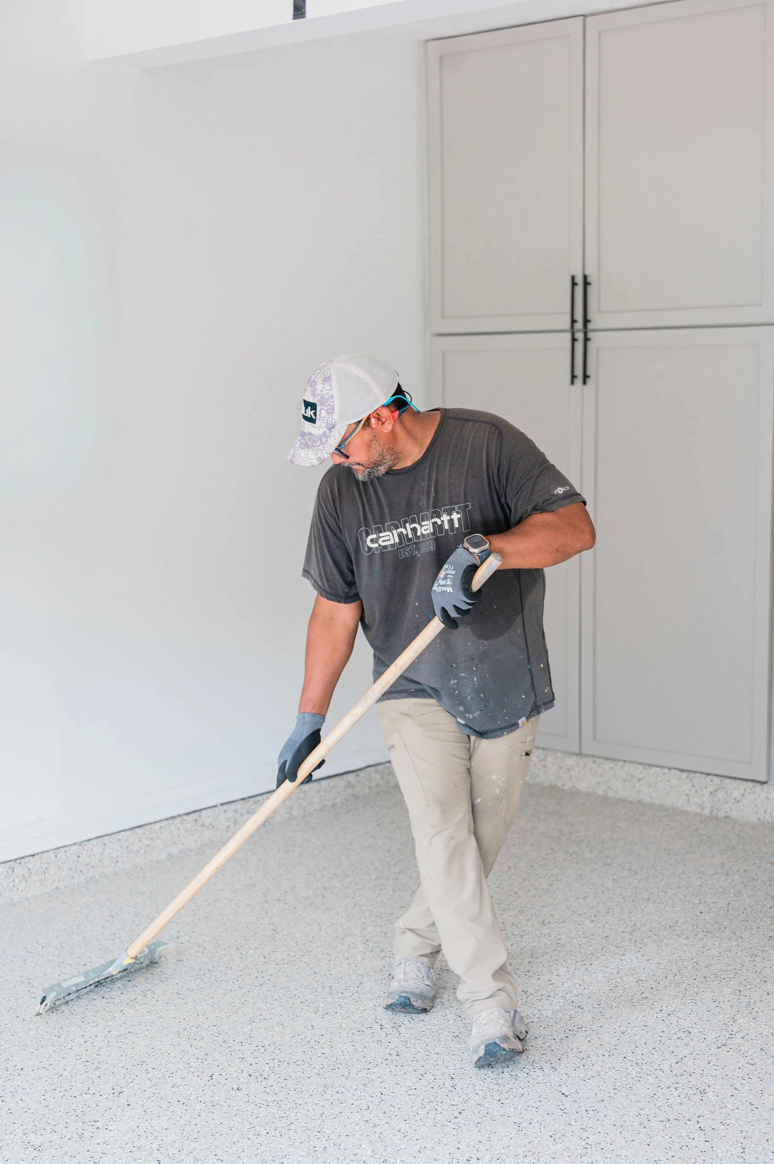 A man wearing a carhartt t-shirt, beige pants, gray shoes, a white cap, ear protection, glasses, and work gloves is spreading a light-colored epoxy or paint on a floor with a long-handled roller in a room with white walls and built-in cabinets.