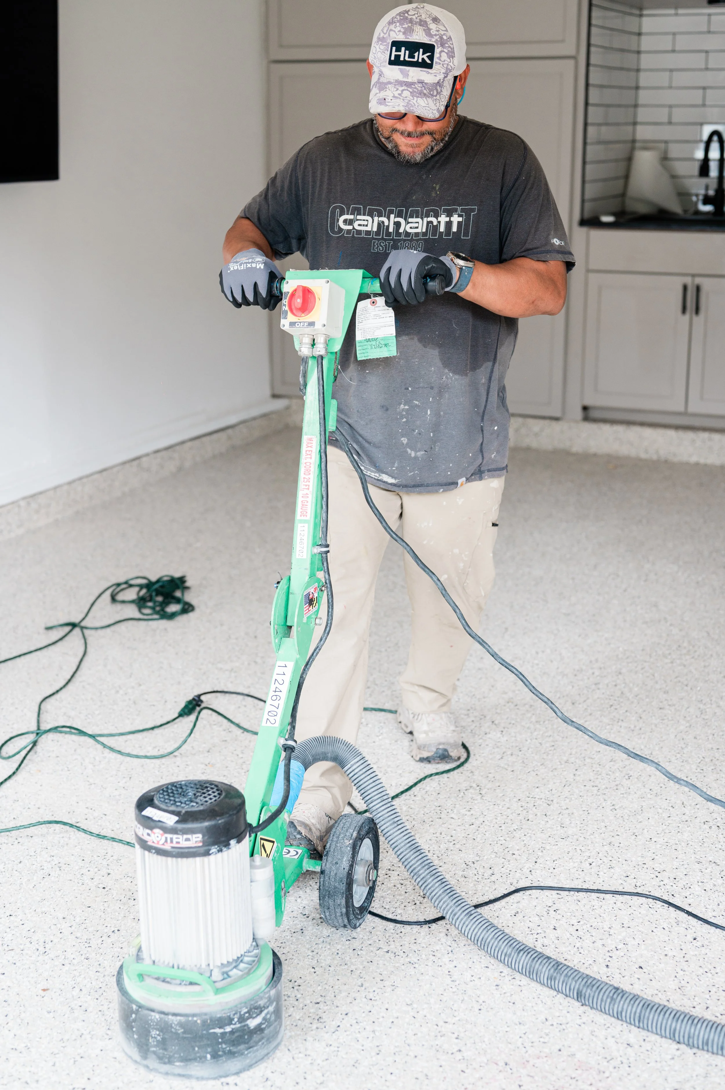 A man operates a green concrete grinder with a large vacuum attachment inside a modern kitchen with light gray cabinets and white tiled walls.
