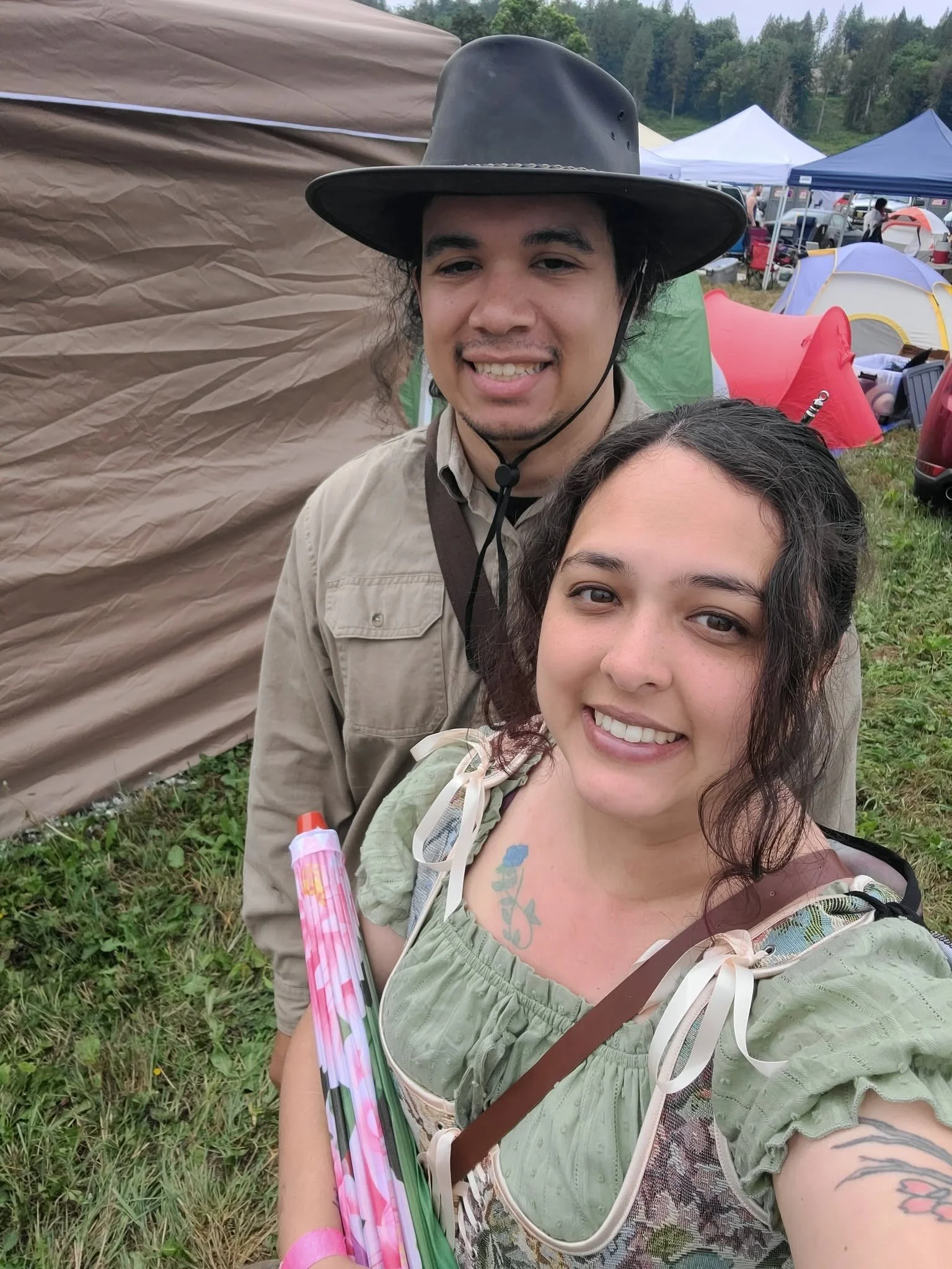 A young woman and man smiling at a grassy outdoor event with tents and trees in the background, the woman holding a pink and green umbrella.
