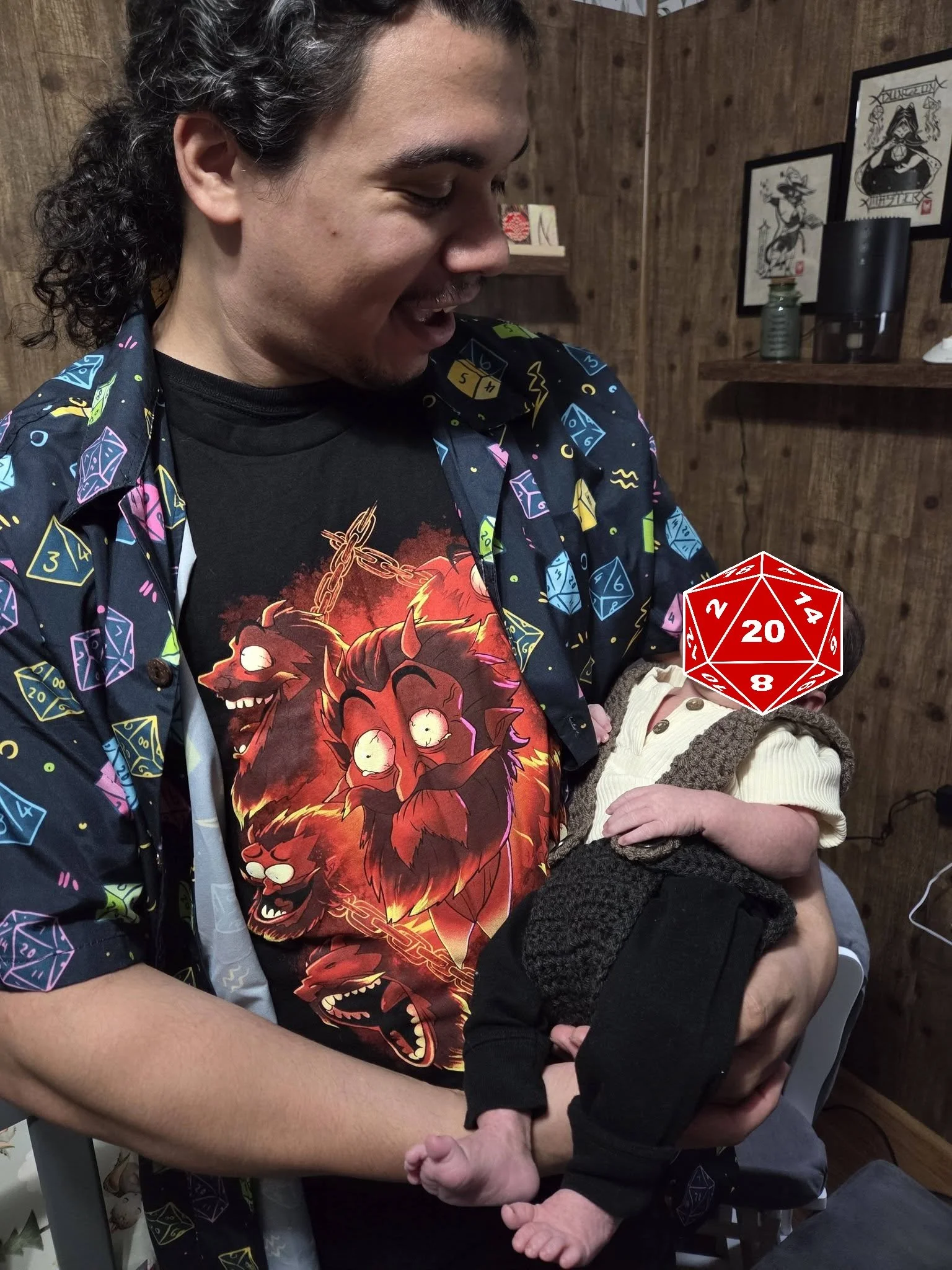 Man holding a baby with a large red 20-sided dice in the foreground, both sitting in a room with wooden walls and framed artwork.