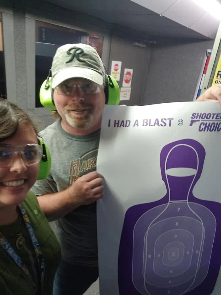 Two people wearing safety glasses and ear protection at a shooting range, holding a target diagram with the text 'I HAD A BLAST' and 'SHOOTER'S CHOICE'.