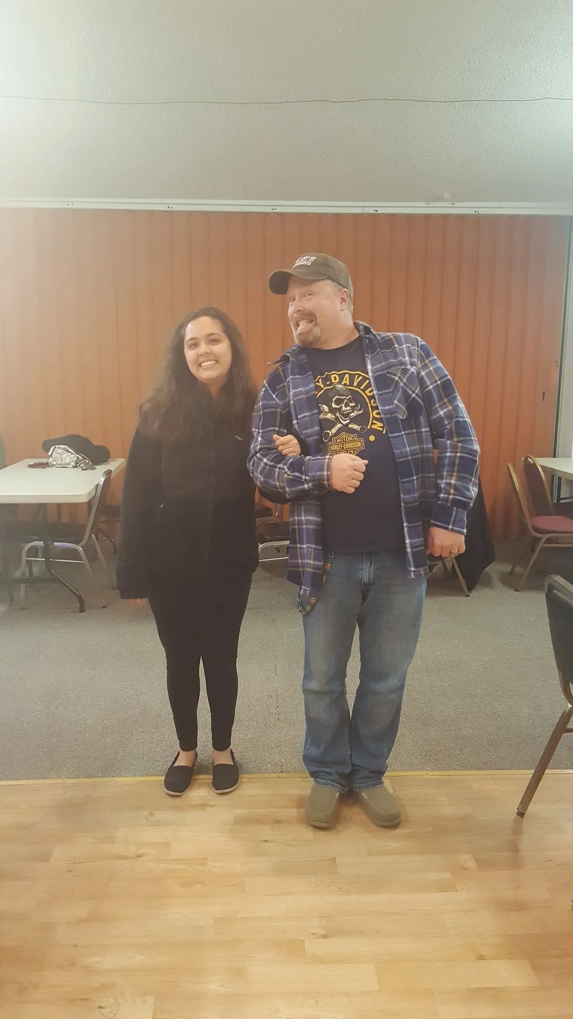 A man and a woman standing indoors, smiling at the camera. The woman has long dark hair, is dressed in black, and is wearing slip-on shoes. The man is wearing a baseball cap, a Harley Davidson t-shirt, a plaid shirt, and jeans. They are standing in front of a wooden wall with tables and chairs in the background.