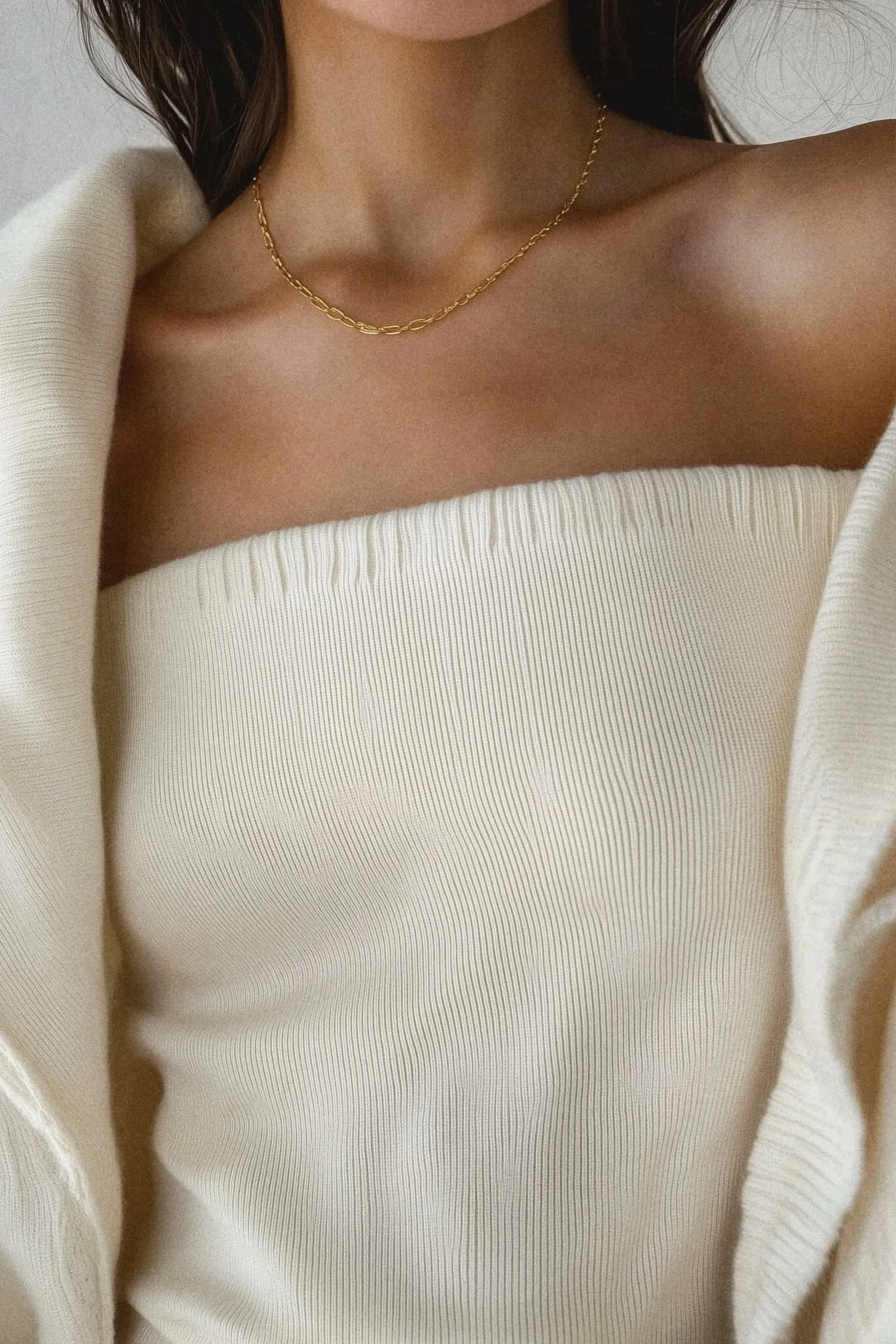 A woman wearing a strapless cream-colored top, a gold chain necklace, and a cream-colored cardigan draped over her shoulders.