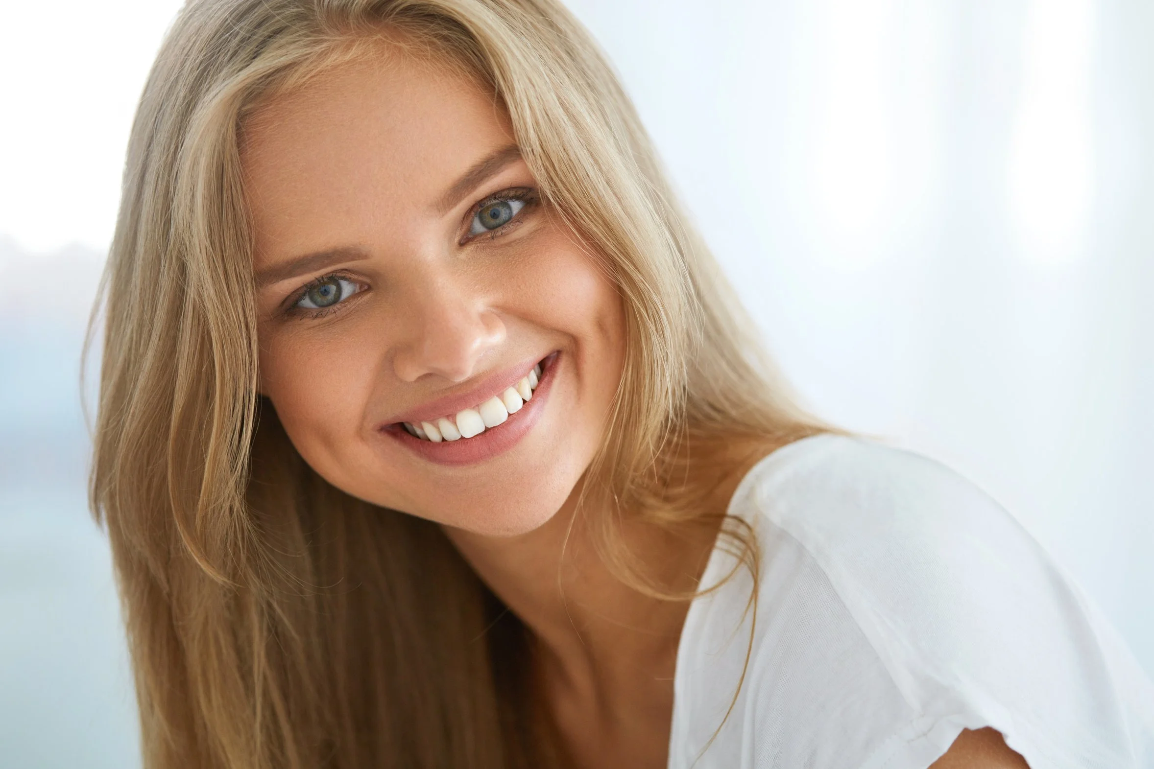 Close-up of a smiling woman with long blonde hair and blue eyes, wearing a white top against a light background.