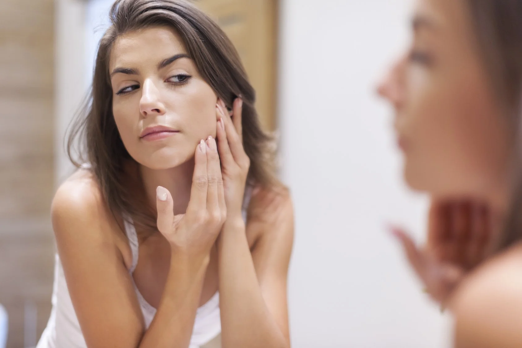 A woman with brown hair looking at herself in the mirror, touching her face with her left hand, appearing to examine her skin.