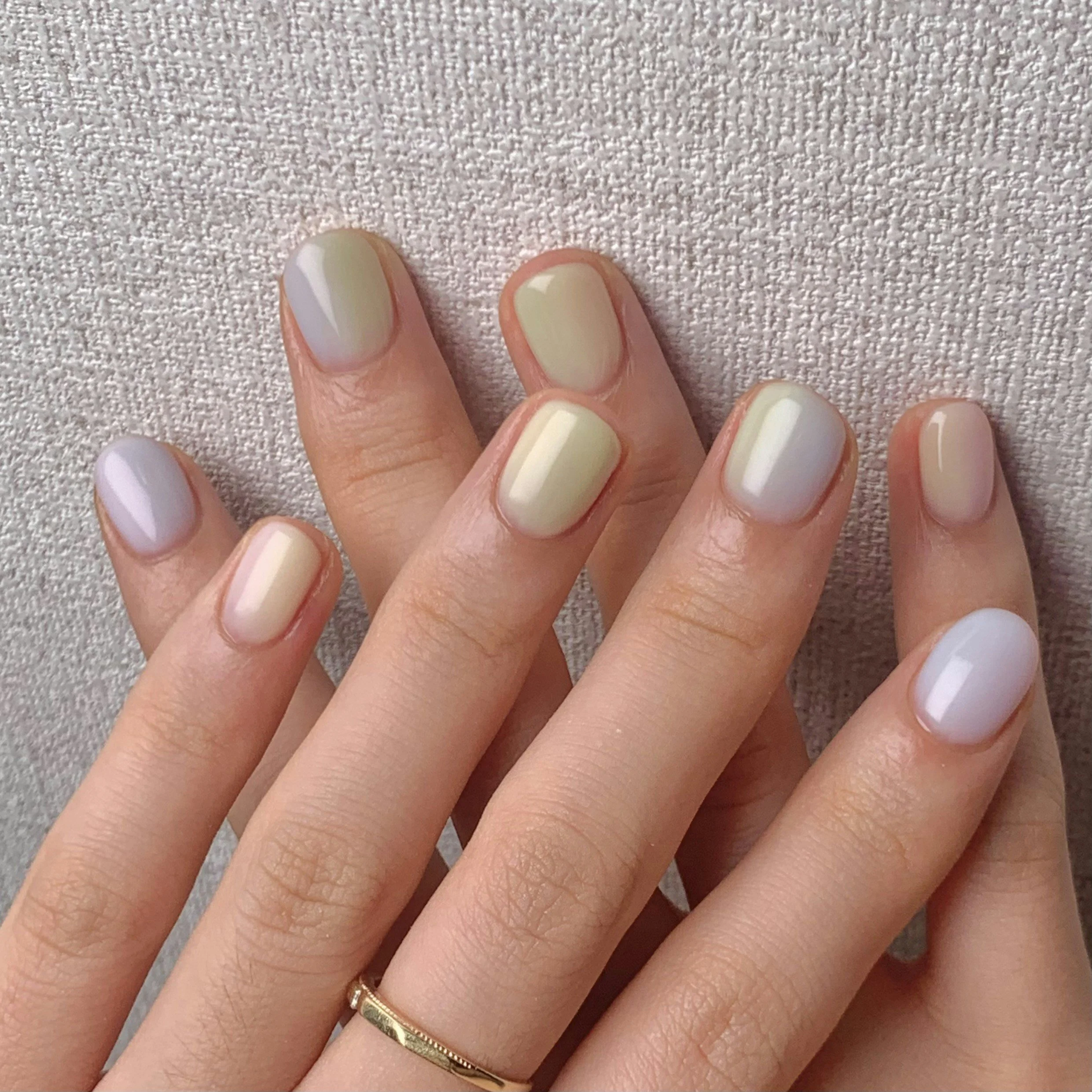 Close-up of a hand with manicured nails painted in a light cream color, wearing two rings, against a textured light-colored fabric background.