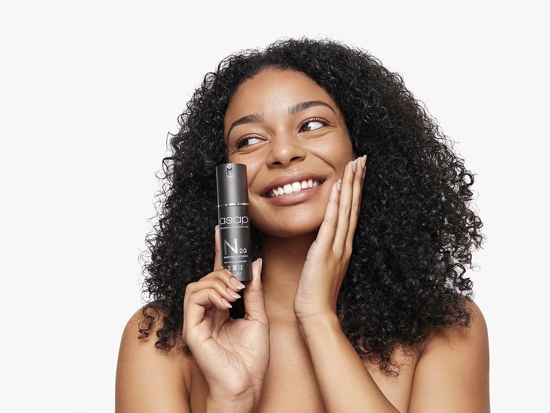 A woman with curly hair smiling and holding a skincare product near her face.