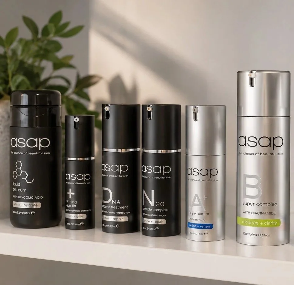 Six skincare bottles from the brand ASAP arranged in a row on a white surface, with a green plant in the background.