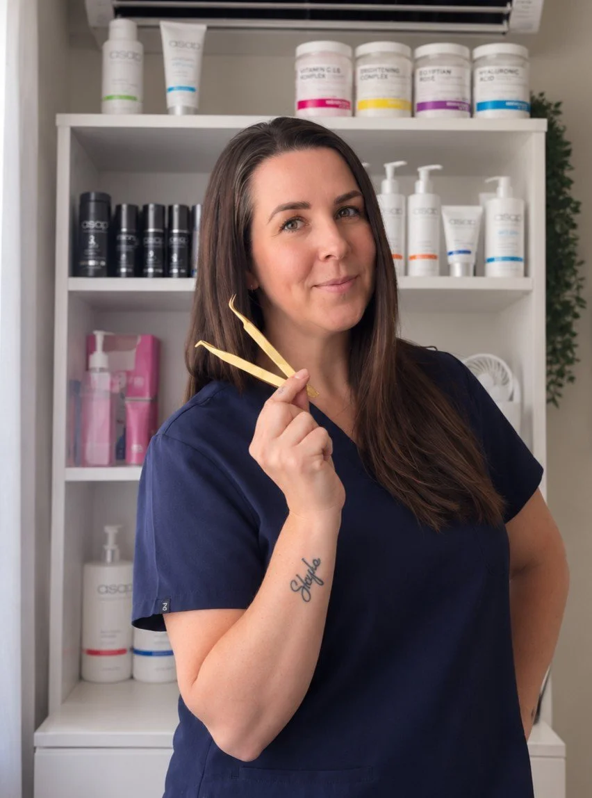 A woman with long brown hair, wearing navy scrubs, standing in front of a white shelf filled with skincare and haircare products, holding two gold-colored tweezers.