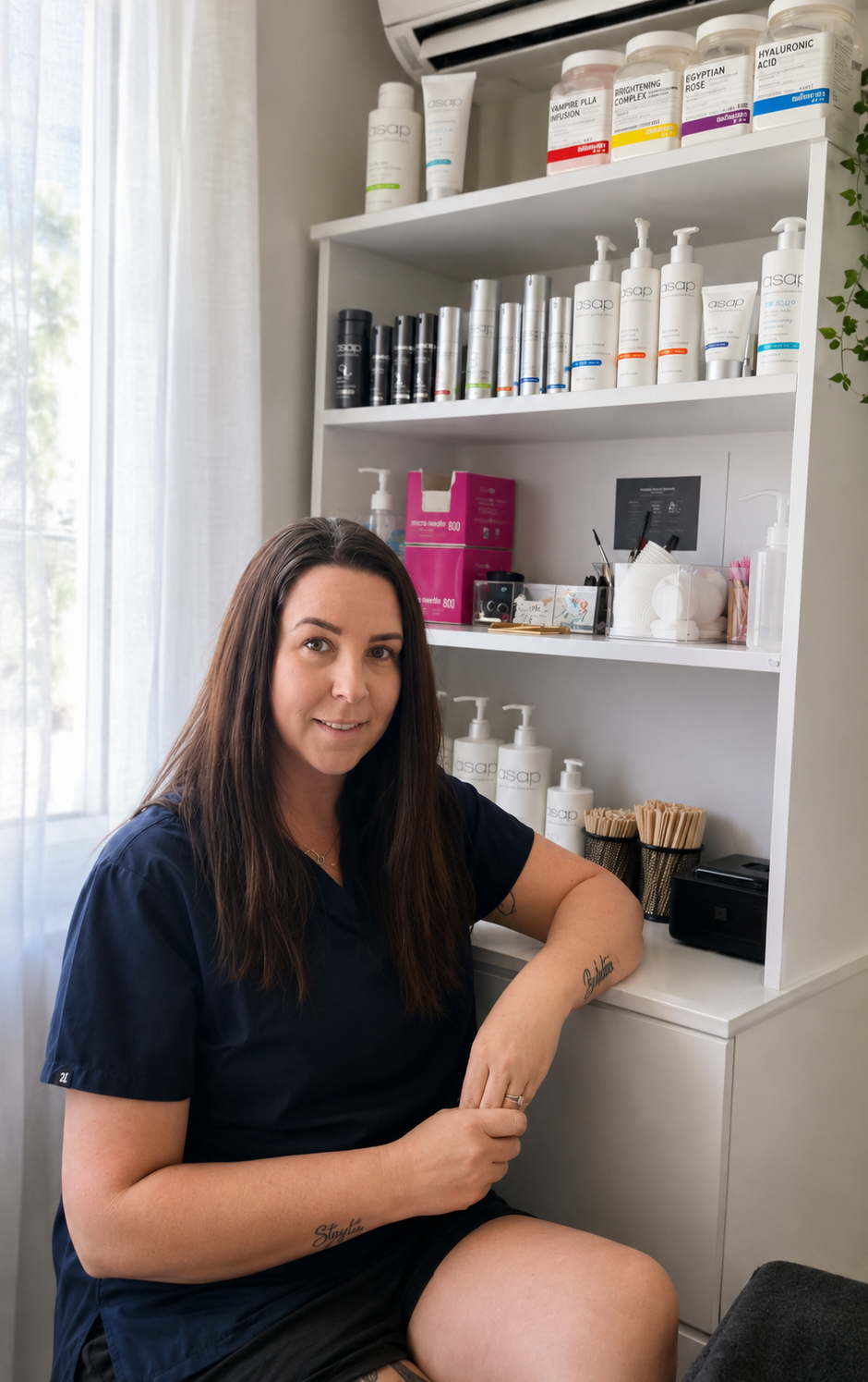 A woman in navy scrubs sitting beside a white shelf containing skincare products, tools, and supplies, inside a clinic or spa with a window and curtain in the background.