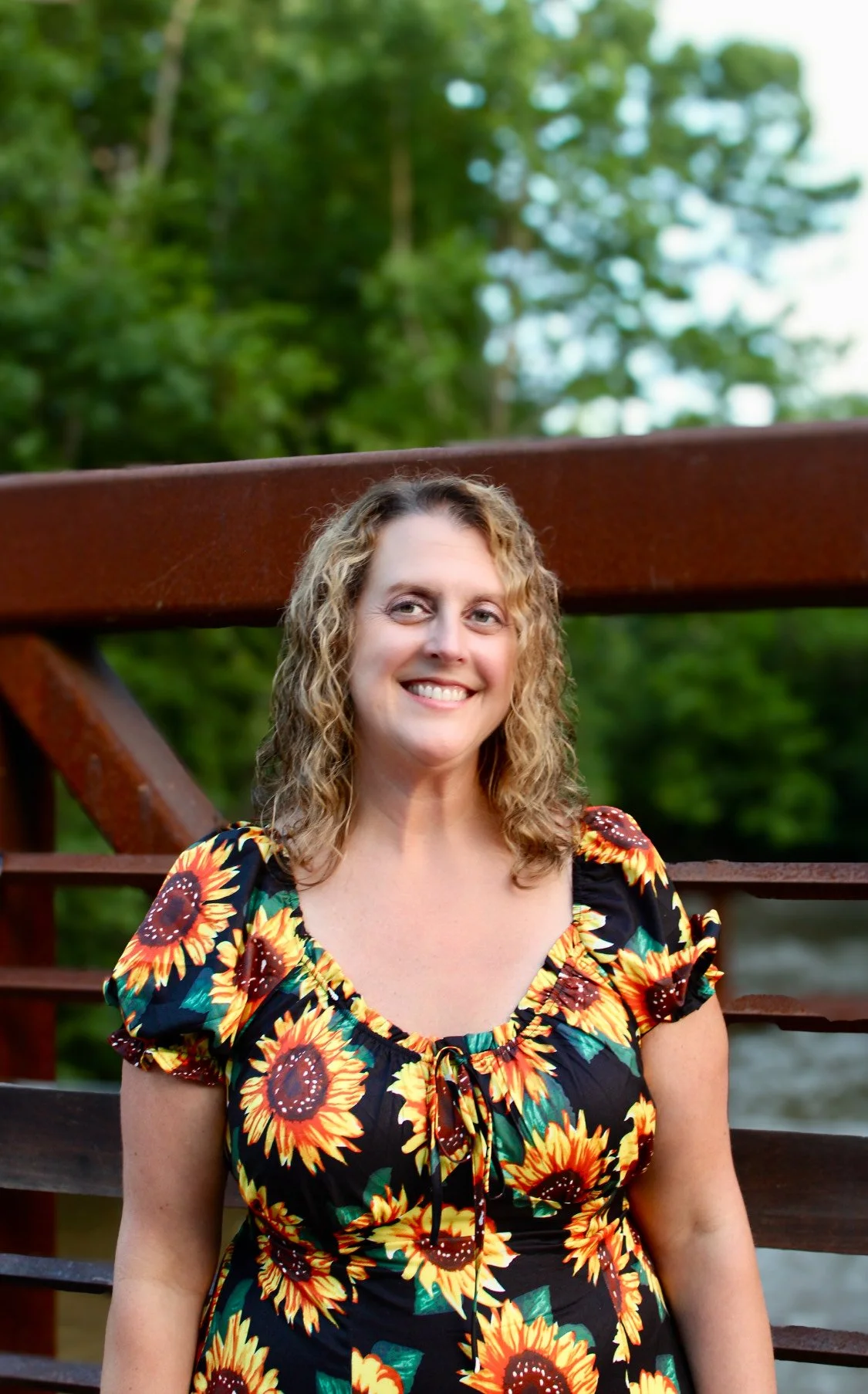 A smiling woman with curly blonde hair wearing a sunflower-patterned dress outdoors with green trees and a brown railing in the background.