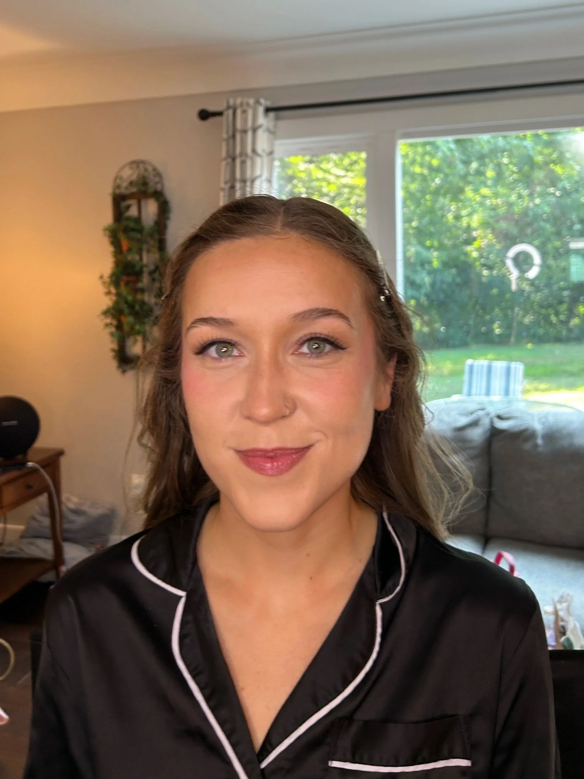Close-up of a woman with light brown hair and green eyes, wearing black pajamas with white piping, smiling subtly. The background shows a cozy living room with a window overlooking a green backyard, a couch, and decorative wall shelves.